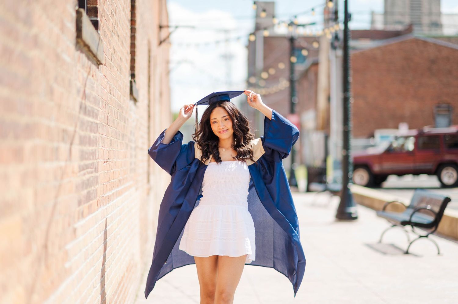 Graduate in navy gown adjusting cap while walking on sunny urban sidewalk.