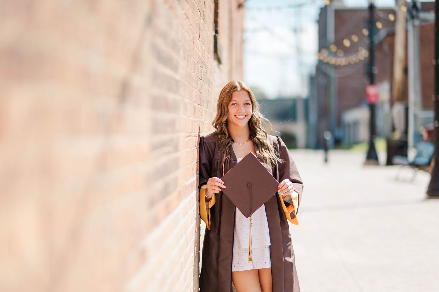 Smiling graduate in brown gown leaning against brick wall holding graduation cap.