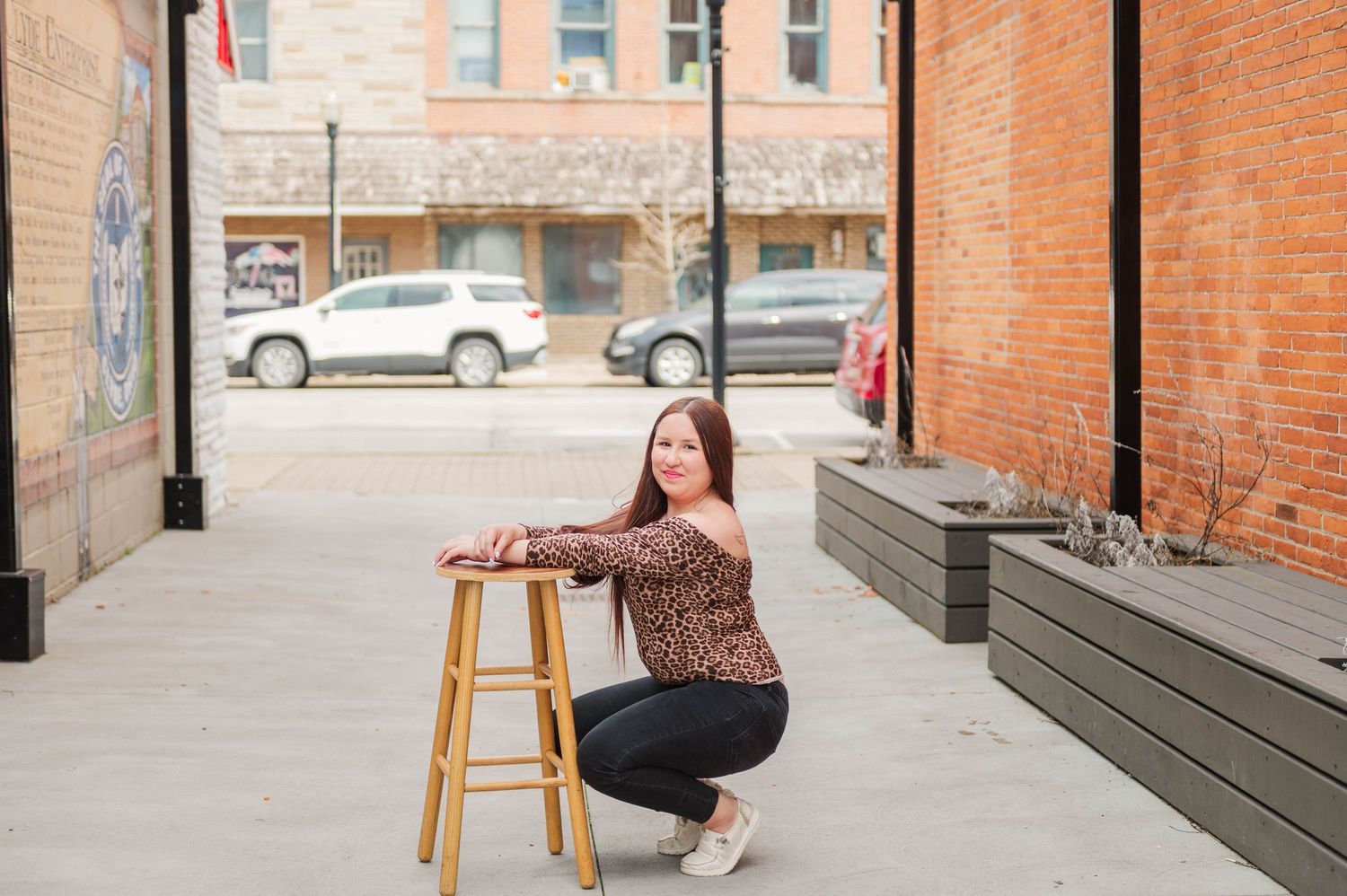 Woman smiling and posing by a wooden stool on a brick-lined urban sidewalk.