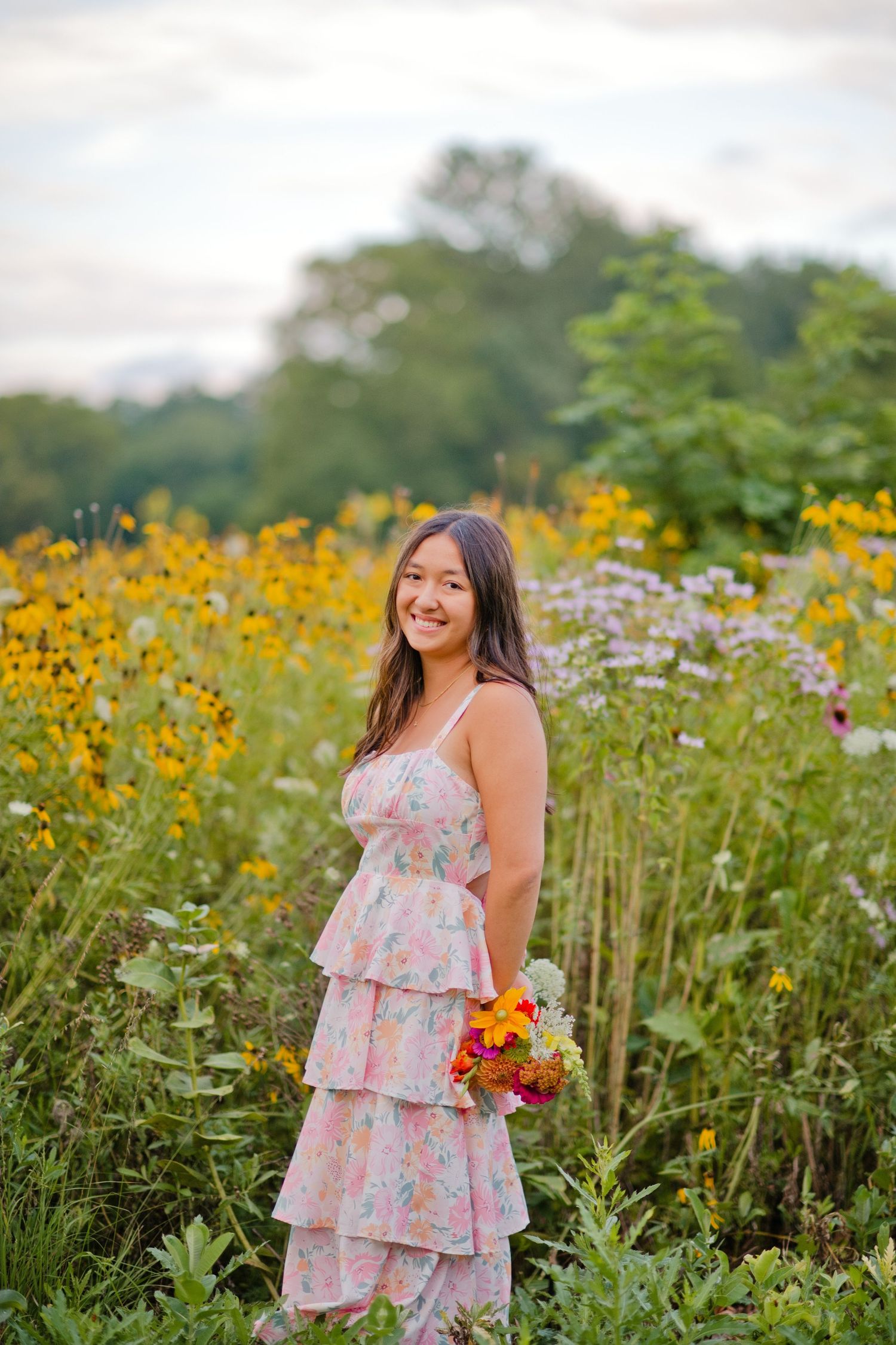 Young woman in floral tiered dress holding bouquet, smiling in vibrant wildflower field with yellow and purple blooms.