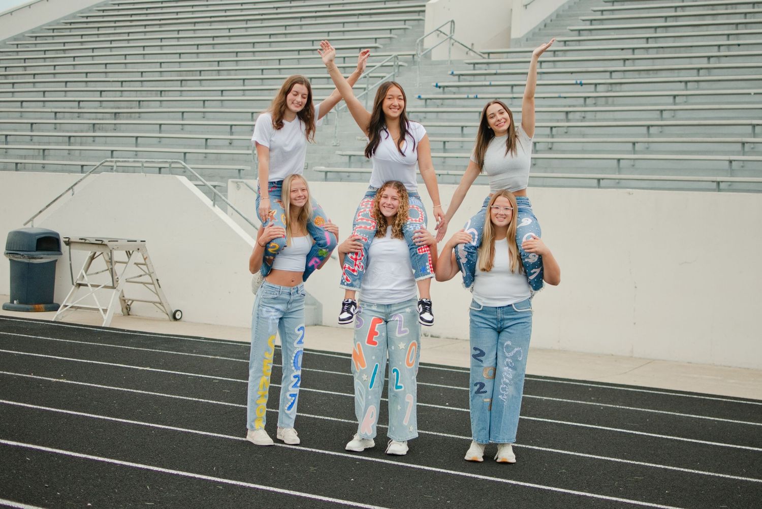 Six teenage girls in white shirts and jeans pose in a pyramid formation on a track field with stadium bleachers behind them.