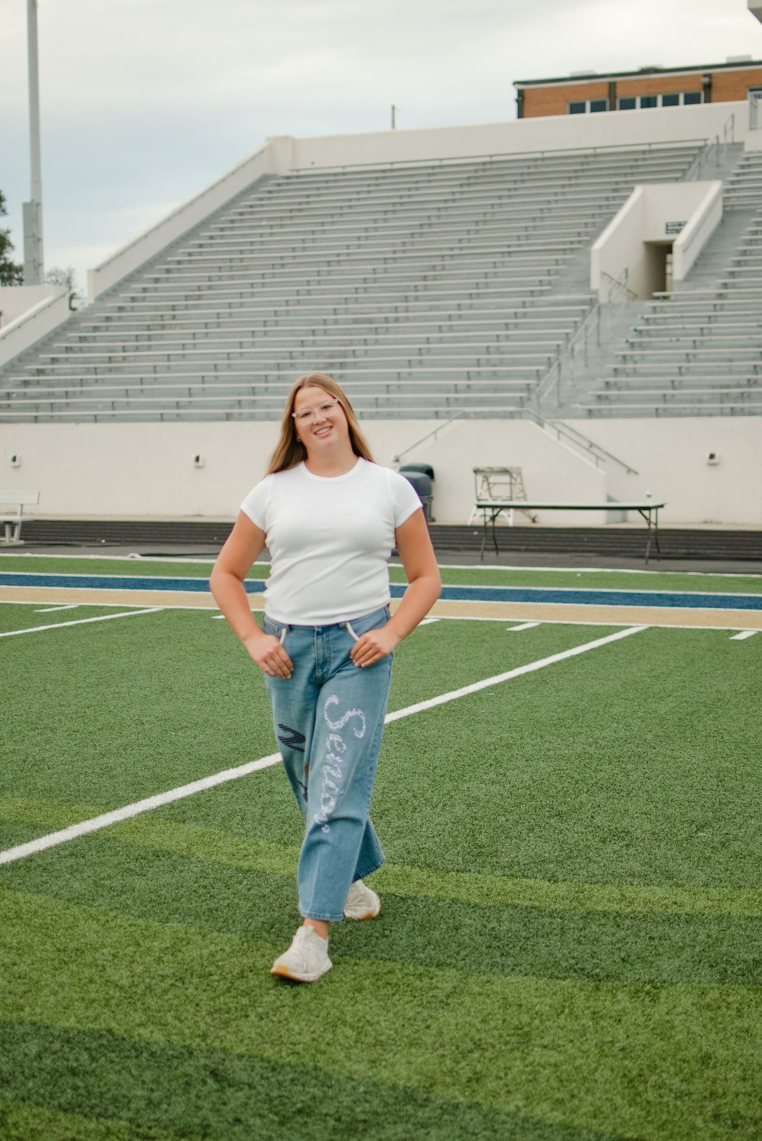 Smiling girl in white tee and painted senior jeans poses confidently on a green football field.