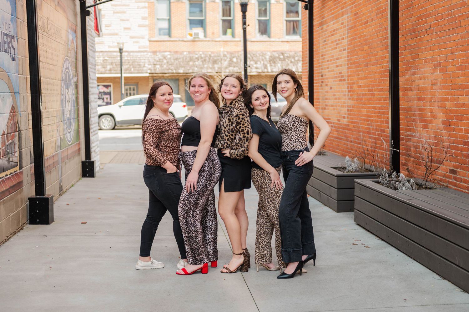 Group of five women in elegant evening wear smiling together in a brick city alley.