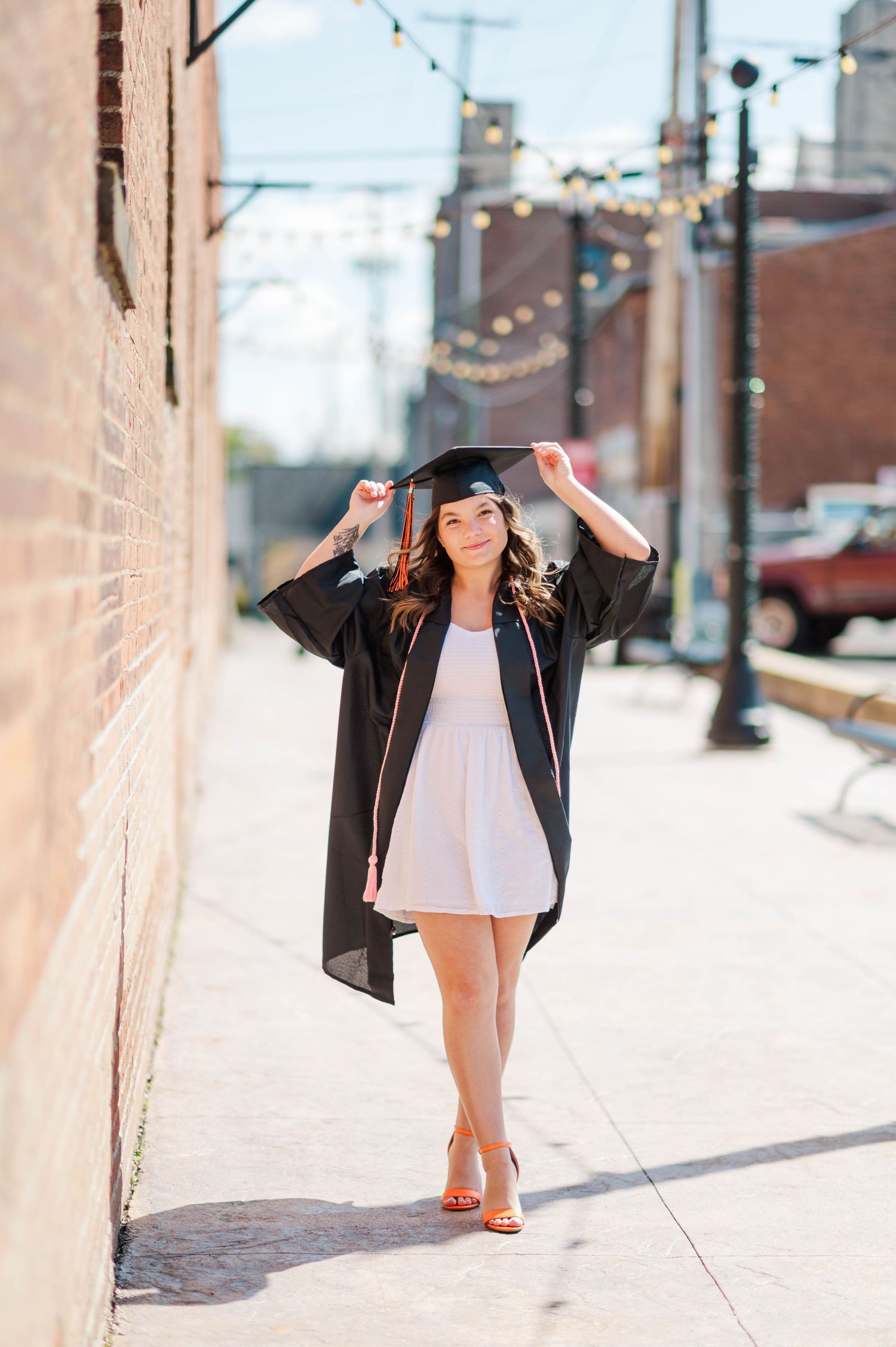 Smiling graduate in black cap and gown walking along brick wall on sunny urban sidewalk.