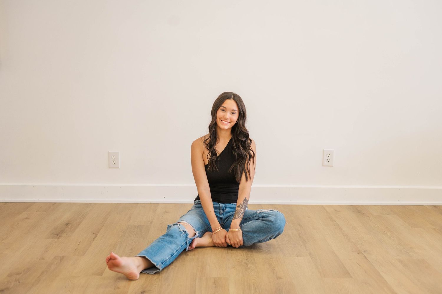 Young woman with long dark hair sitting cross-legged on hardwood floor wearing black top and ripped jeans.