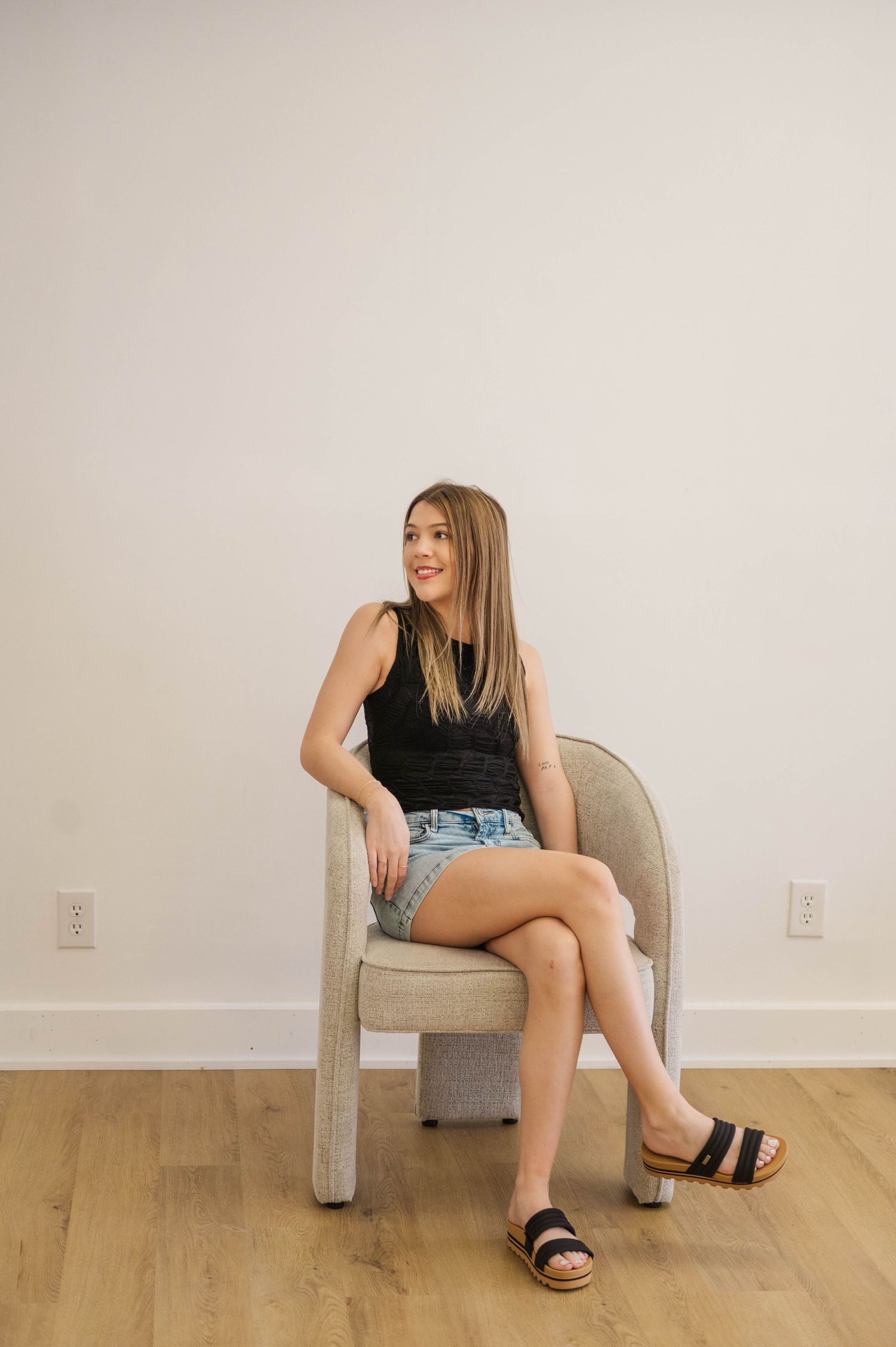 Young woman in black tank top and denim shorts sitting on a modern chair in a bright minimalist room.