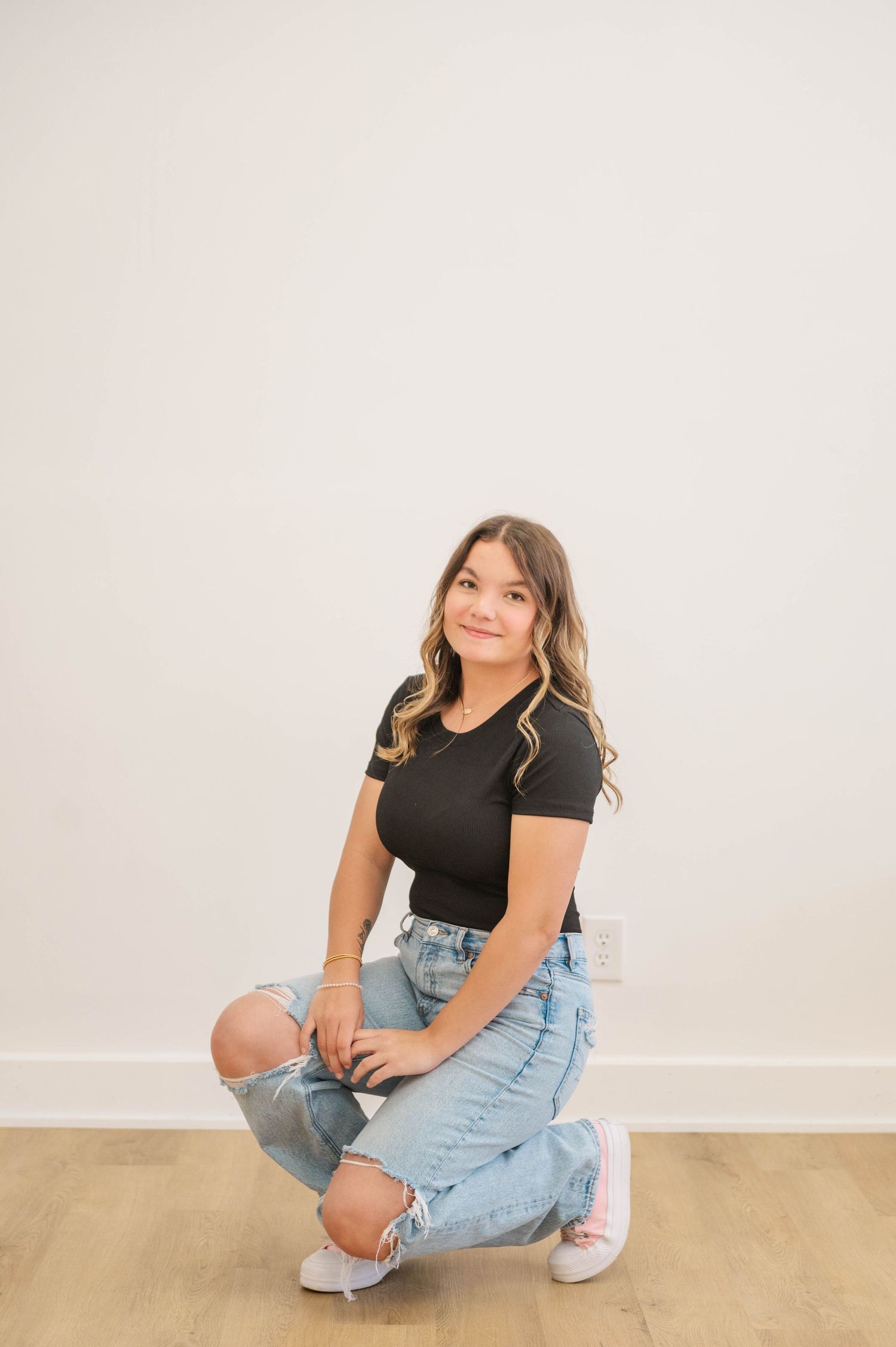 Young woman in black tee and ripped jeans kneeling on wood floor against white wall.