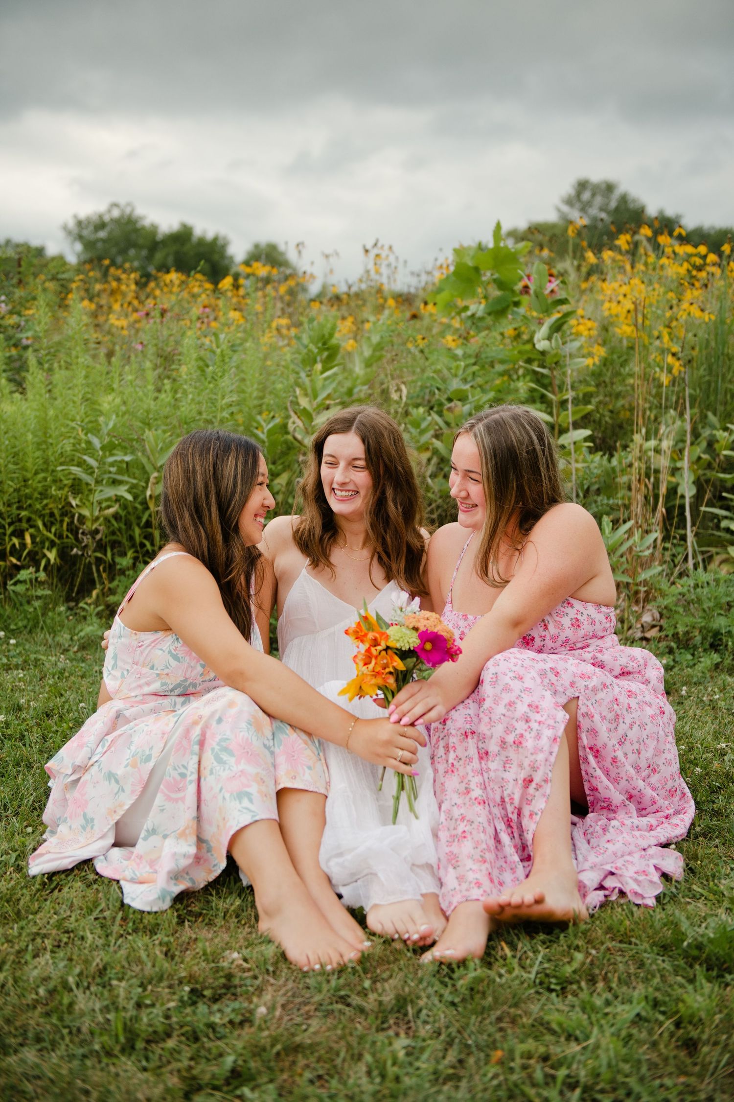 Three young women in floral dresses sit barefoot in a wildflower field laughing and sharing a bouquet.