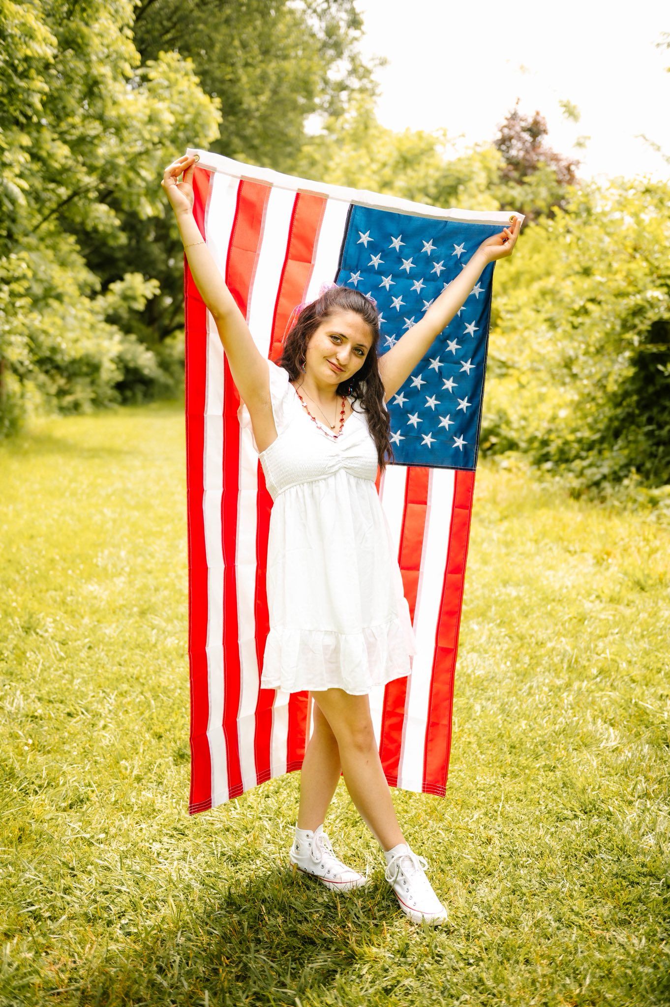 Person in white dress poses joyfully with American flag in sunny meadow.