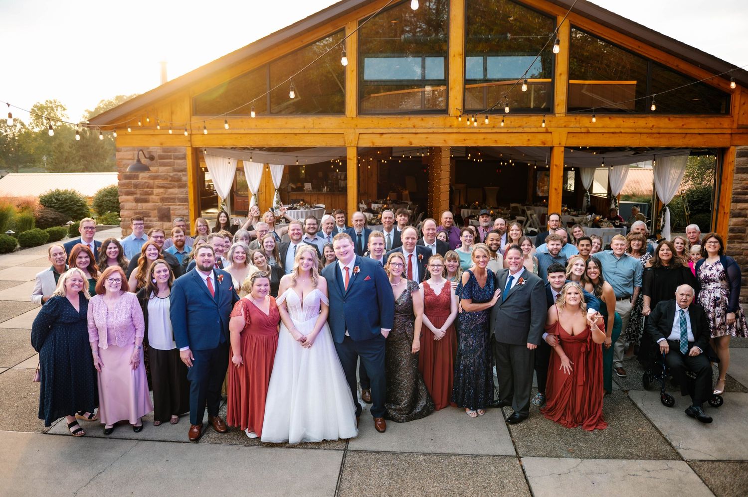 Large wedding party gathered outside rustic venue for group photo.