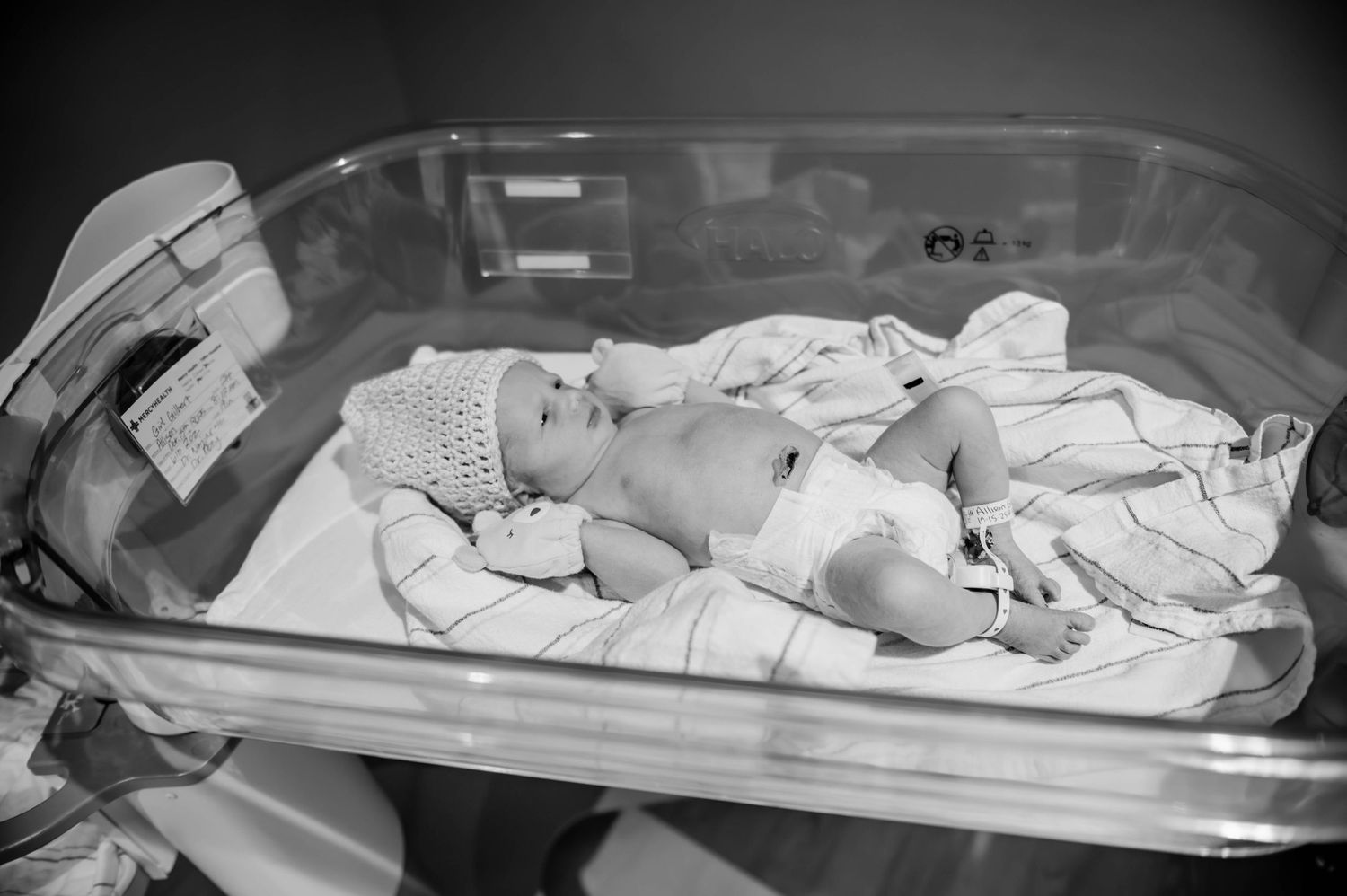 A newborn baby rests in a hospital bassinet wrapped in white blankets and medical monitoring equipment.