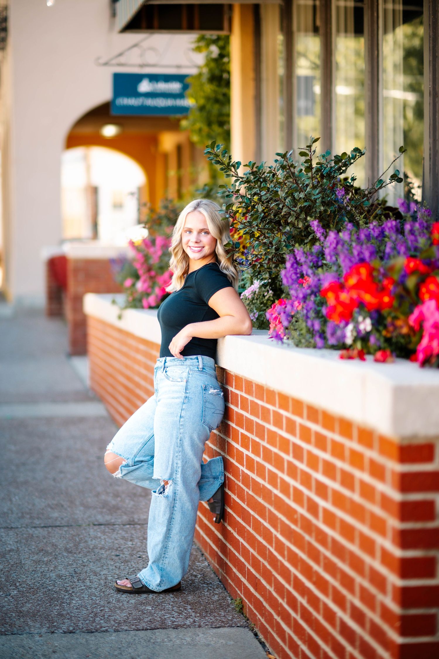 A person in overalls sits on a brick planter with vibrant purple and red flowers along a storefront walkway.