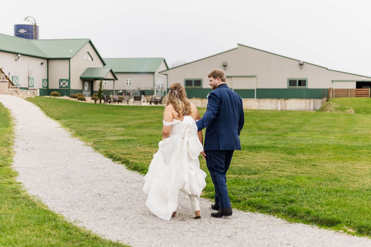 A couple kneels on a gravel path surrounded by green grass and farm buildings.