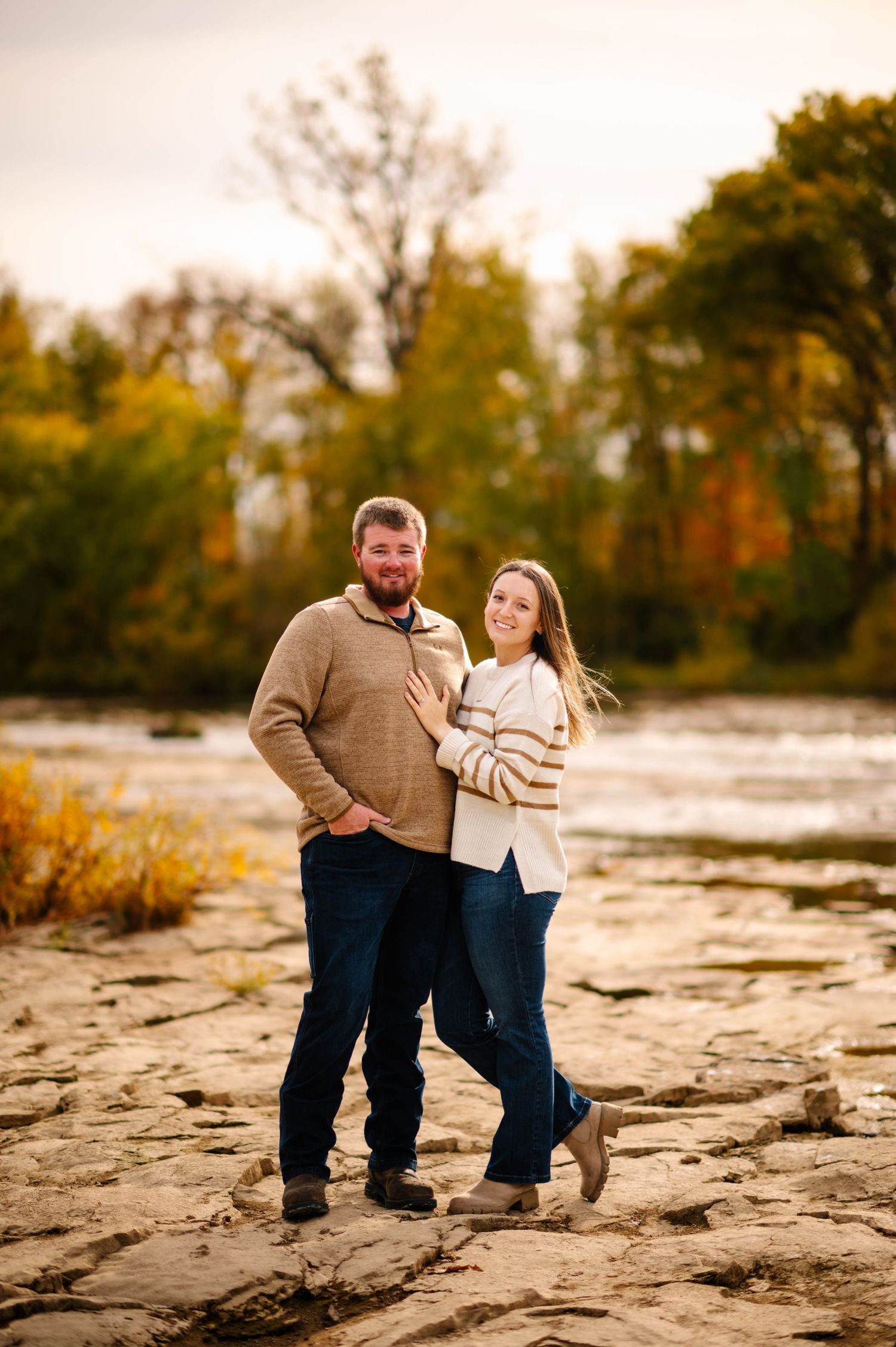 Couple in casual wear stands together on rocky terrain with autumn scenery.