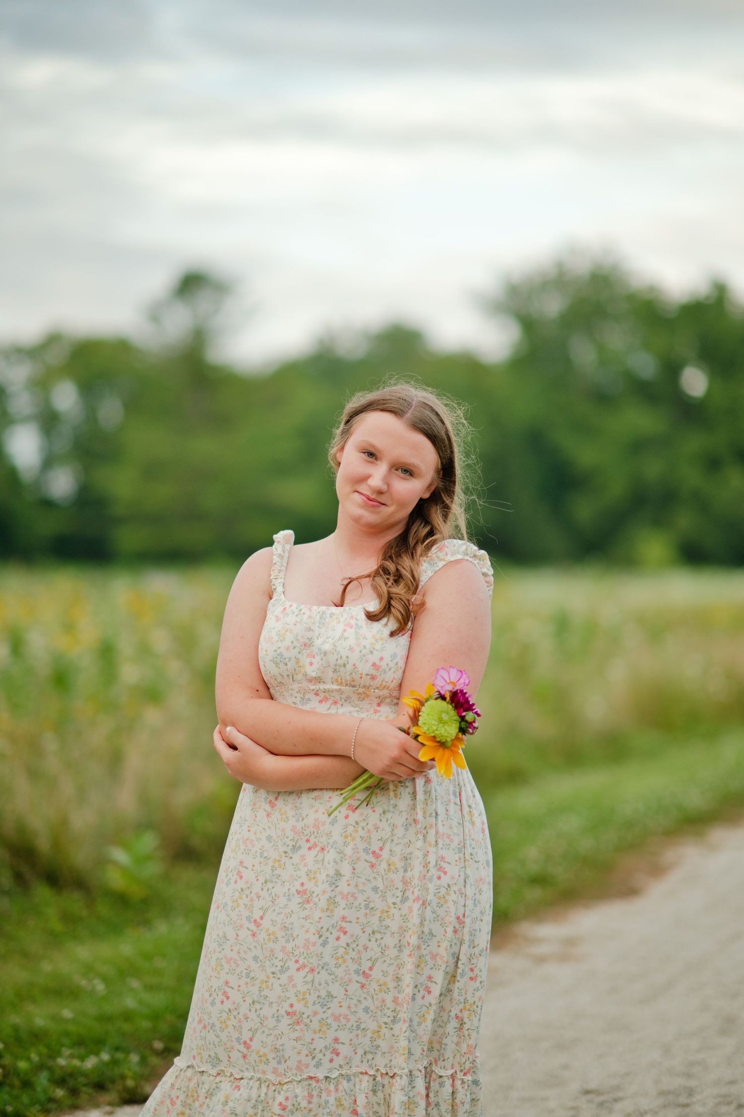 Young woman in a floral sundress holds a colorful bouquet while posing on a path beside a flower field.