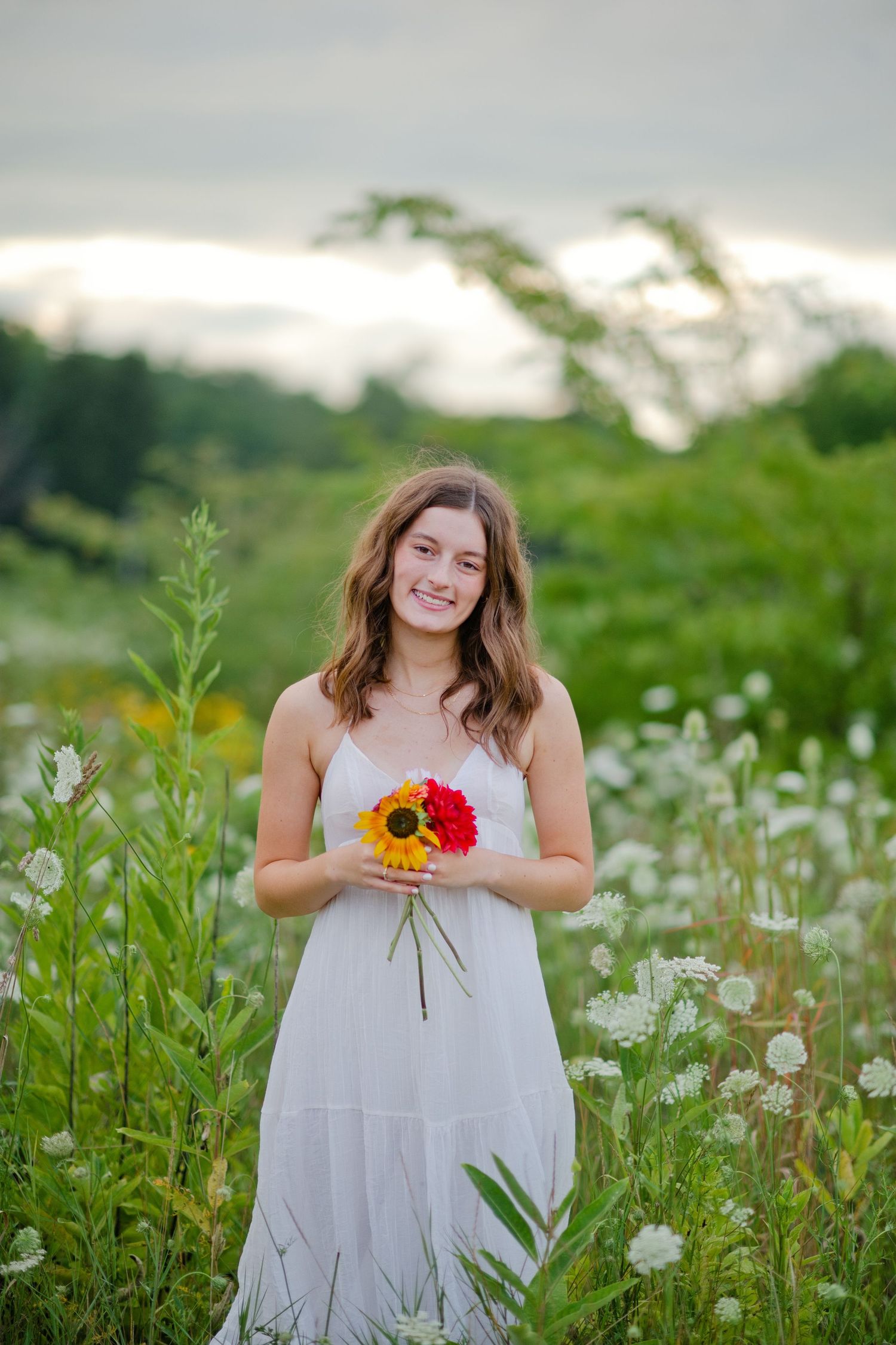 A smiling teenage girl in a white dress holds yellow and red sunflowers in a blooming wildflower field.