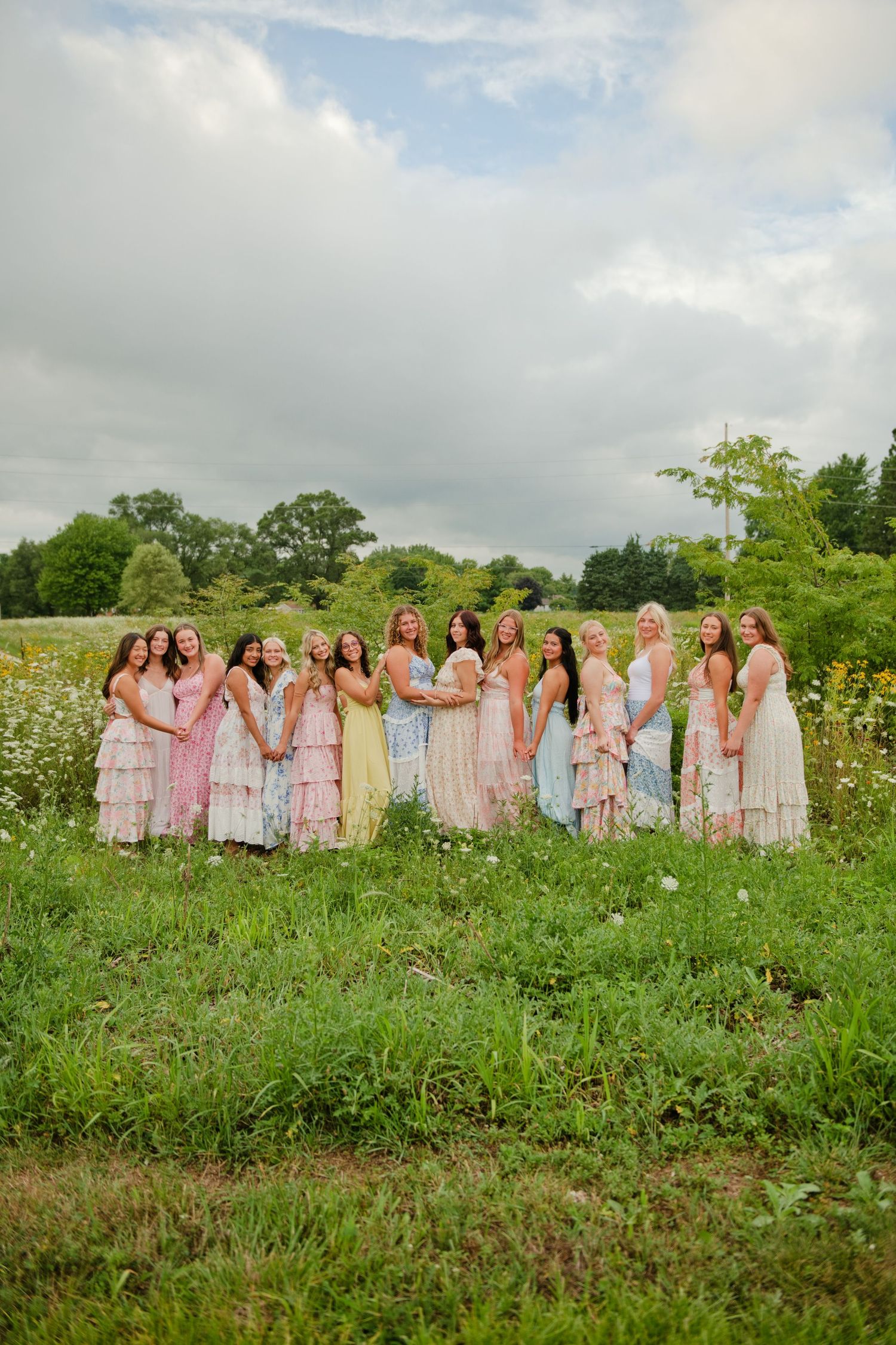 Bridesmaids in pastel dresses gathered together smiling in a lush green field.