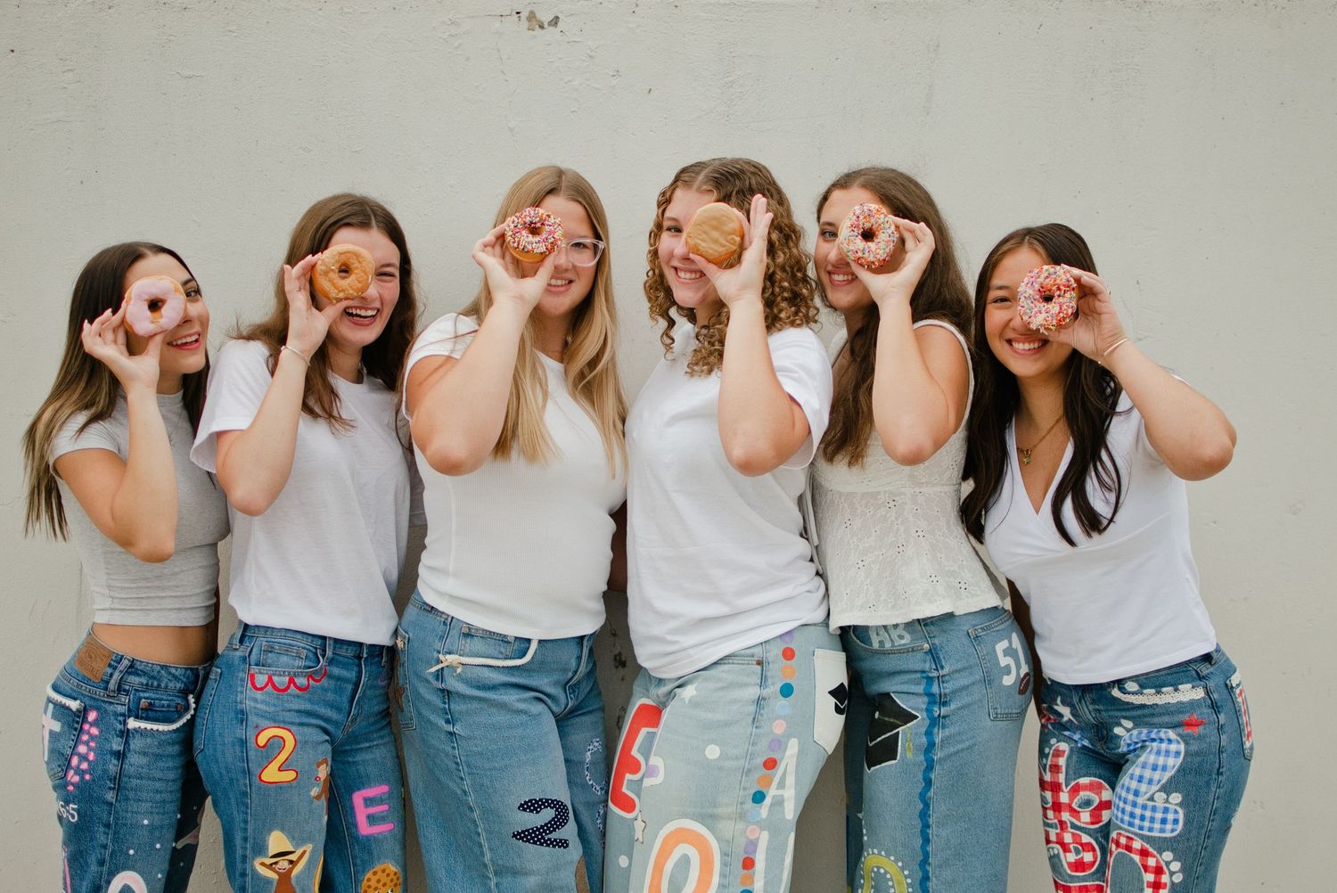 Six girls in white tops and colorful custom jeans hold donuts over their eyes playfully.