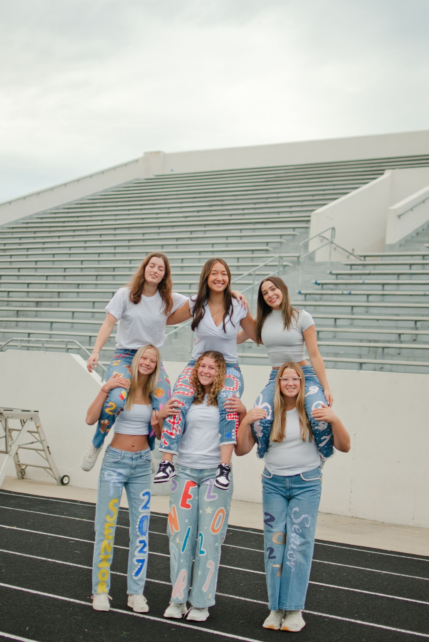 Six teenage girls in white shirts and painted jeans form a fun pyramid pose on a track in front of stadium bleachers.