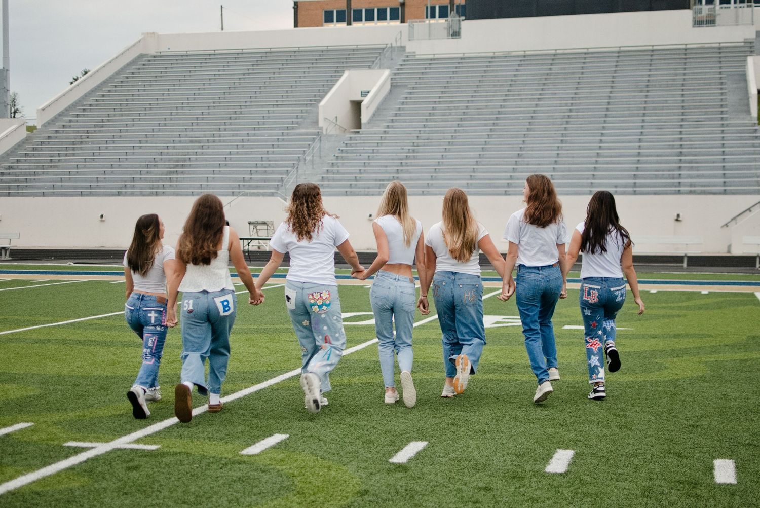 Seven girls in white shirts and jeans walk together on a football field, viewed from behind.