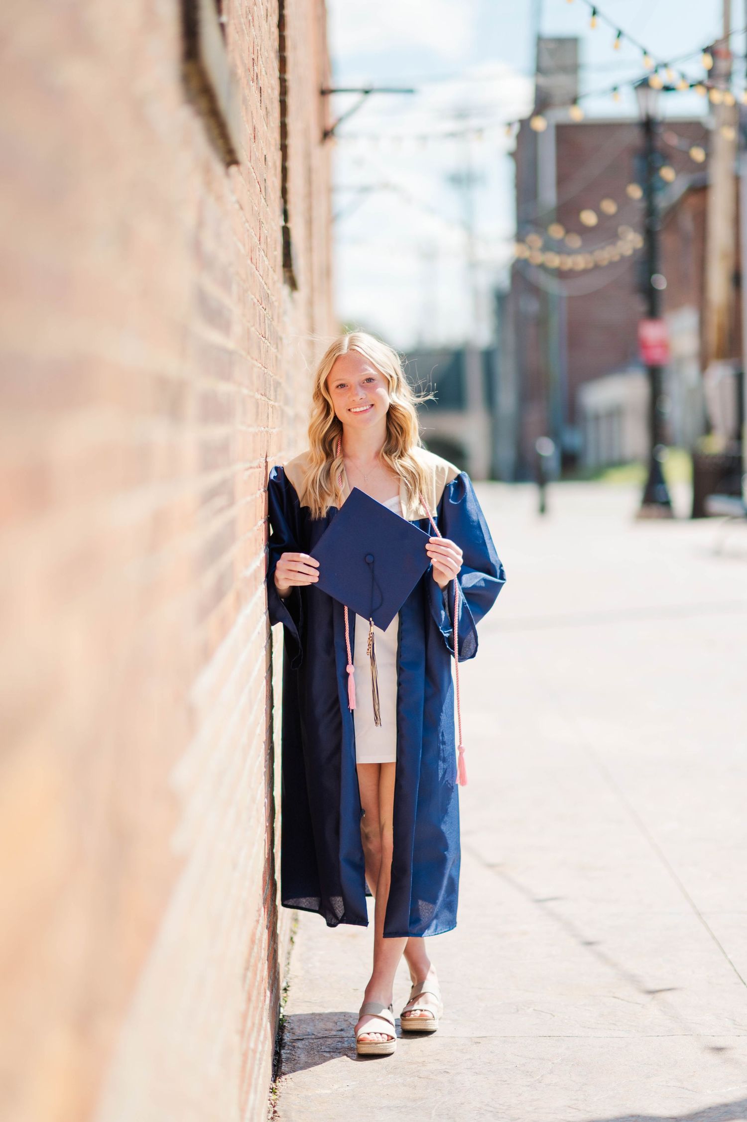 Blonde graduate in navy cap and gown leaning against brick wall on sunny sidewalk.