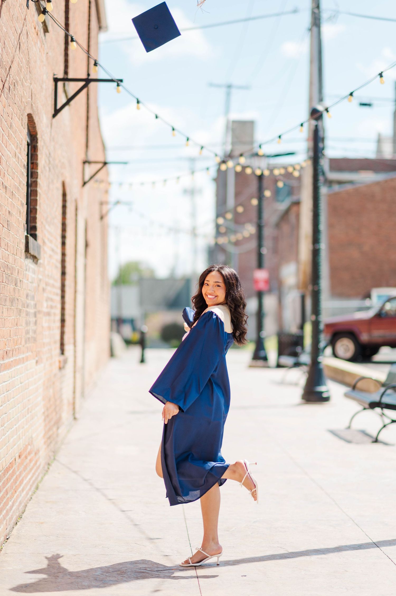 A smiling graduate in a blue cap and gown tosses her mortarboard in the air on a charming brick-lined urban sidewalk.