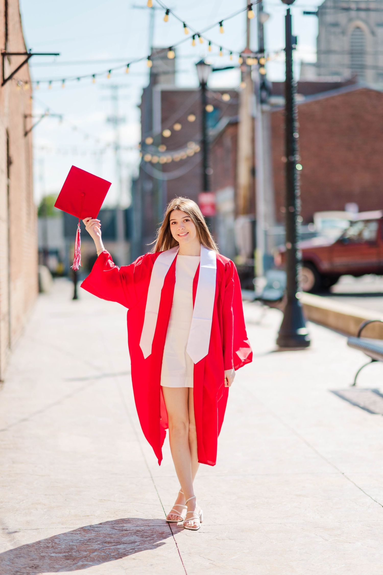 Young woman in red graduation gown holding cap on sunny urban sidewalk.