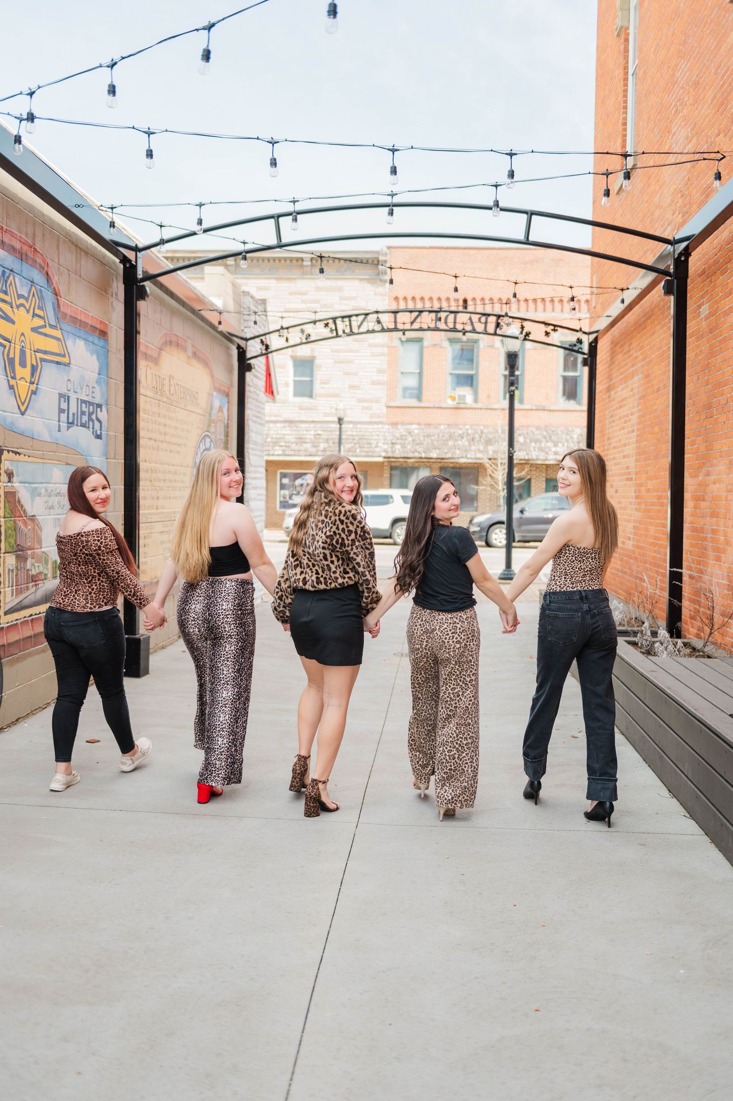 Five young women in leopard print outfits walk hand in hand away from camera under string lights.