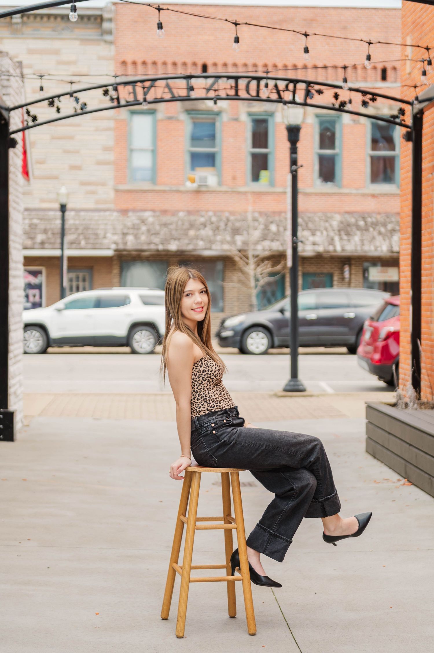 Young woman in leopard top and black jeans sits on wooden stool outdoors in small town setting.