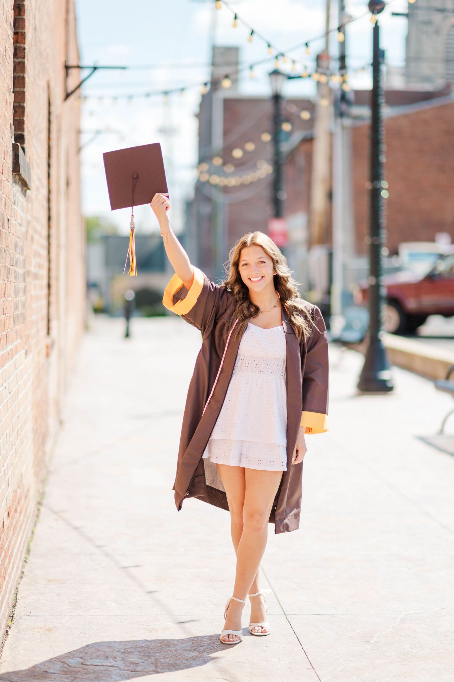 Happy graduate in brown cap and gown holding mortarboard up while walking on sunny brick-lined sidewalk.