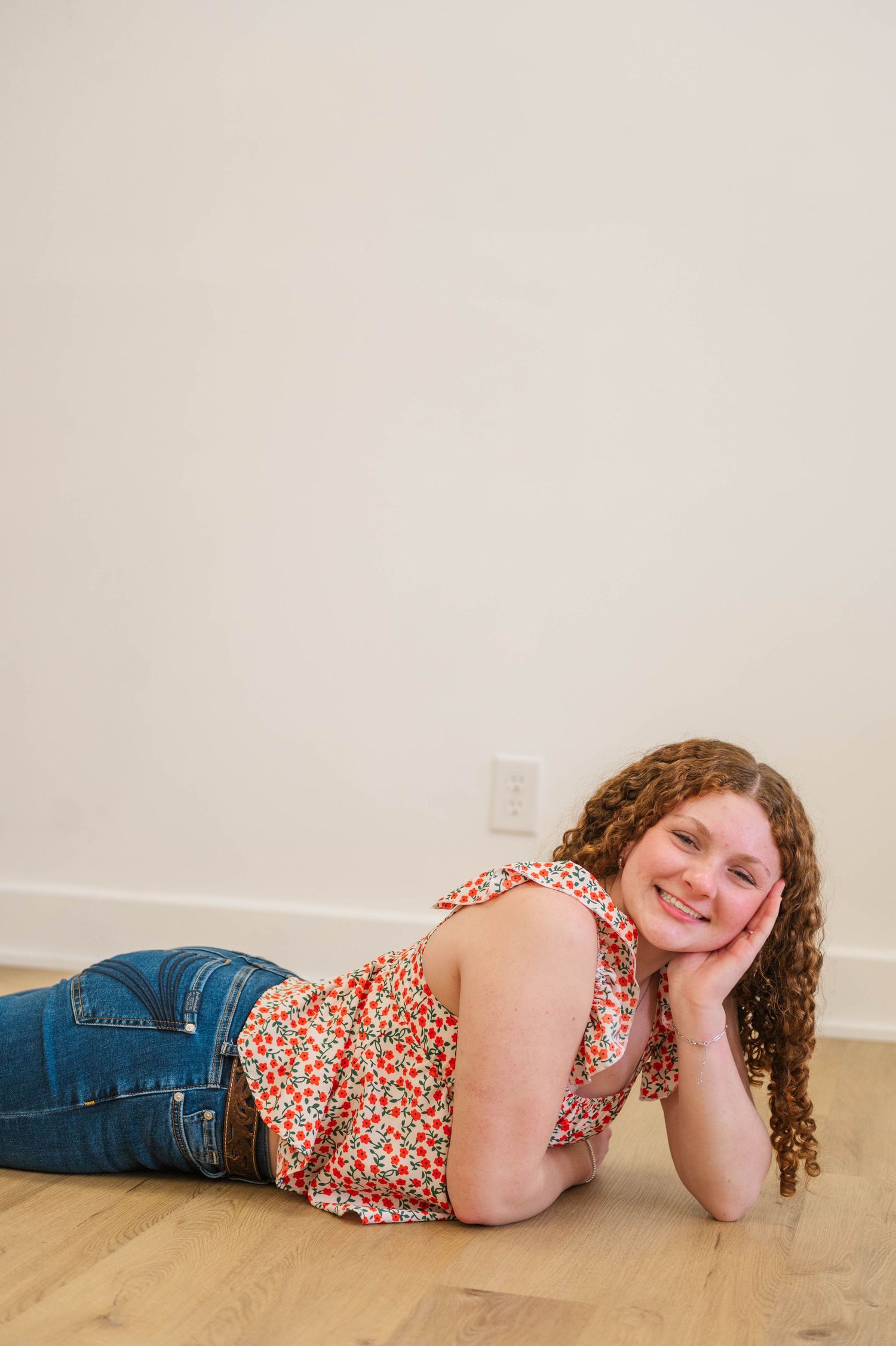 Curly-haired girl in floral top and jeans lying on floor smiling with head resting on hand.
