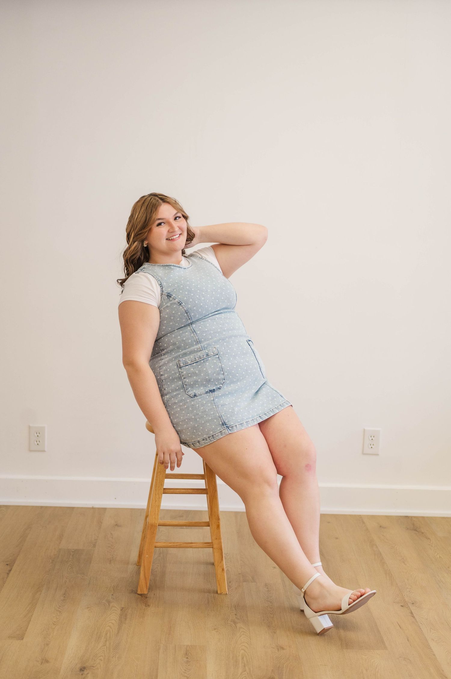 Smiling woman sits on wooden stool in denim overall dress and white tee, hand behind head, wearing sandals.