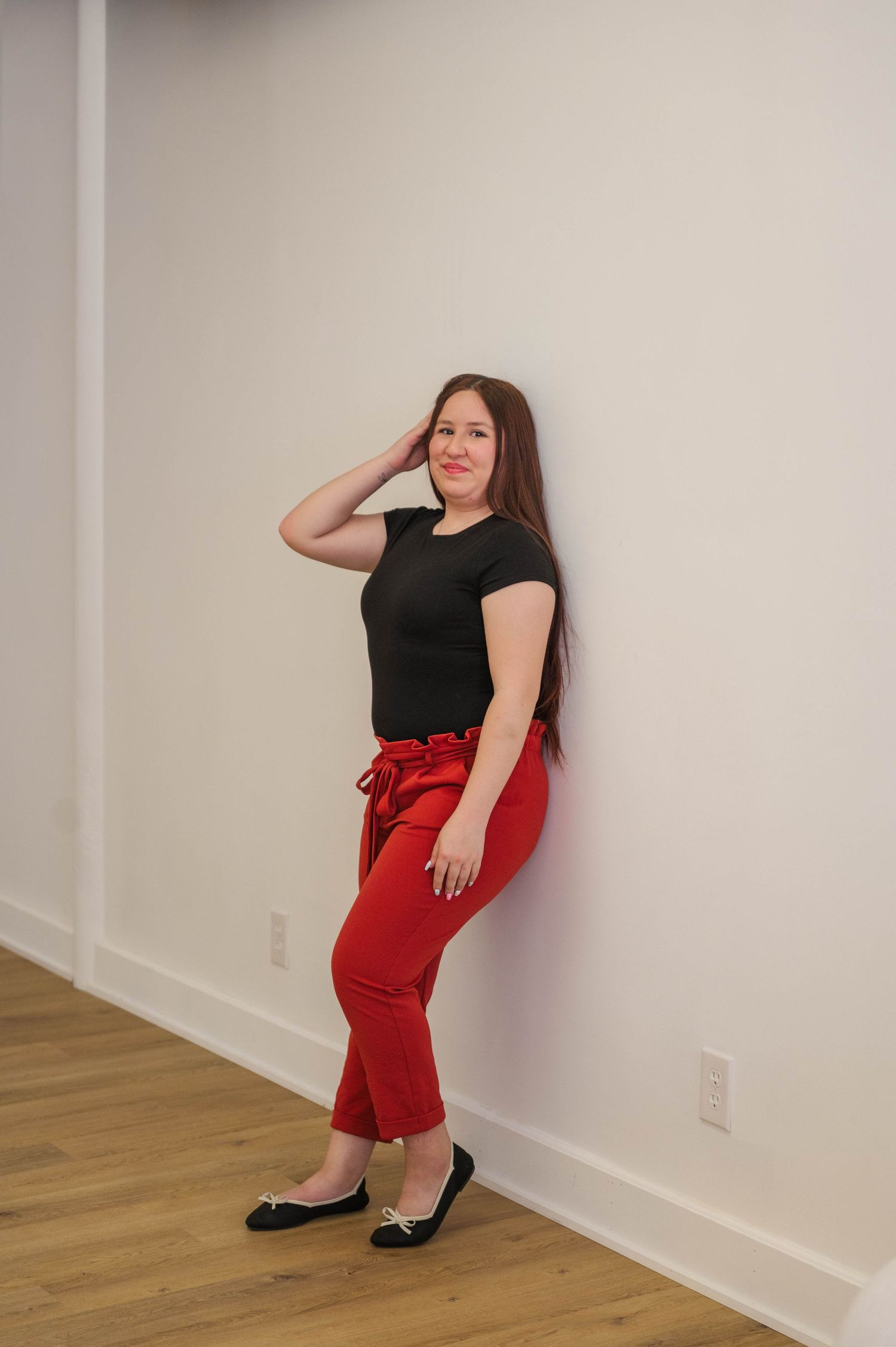 Young woman in black tee and red pants leaning against white wall on hardwood floor.