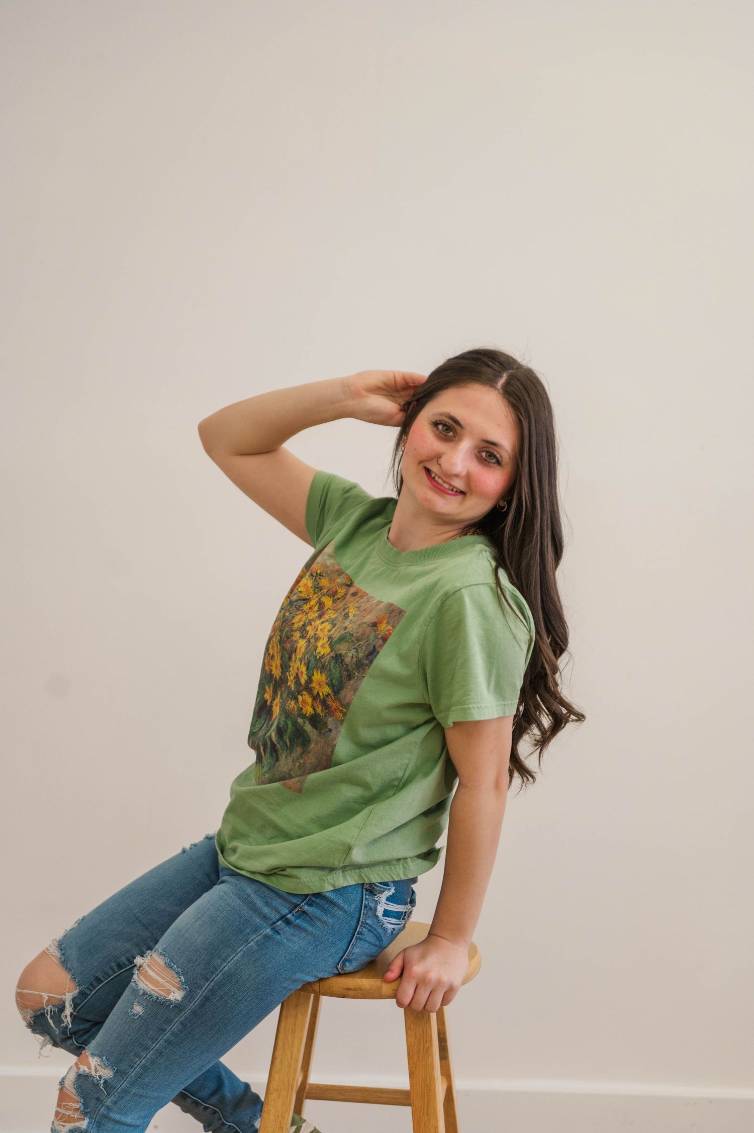 Smiling young woman in green graphic tee and ripped jeans sitting on wooden stool against white background.