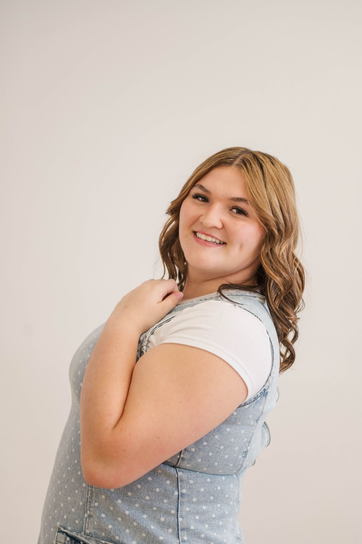 Smiling young woman in white tee and blue polka dot jumper posing against a light background.