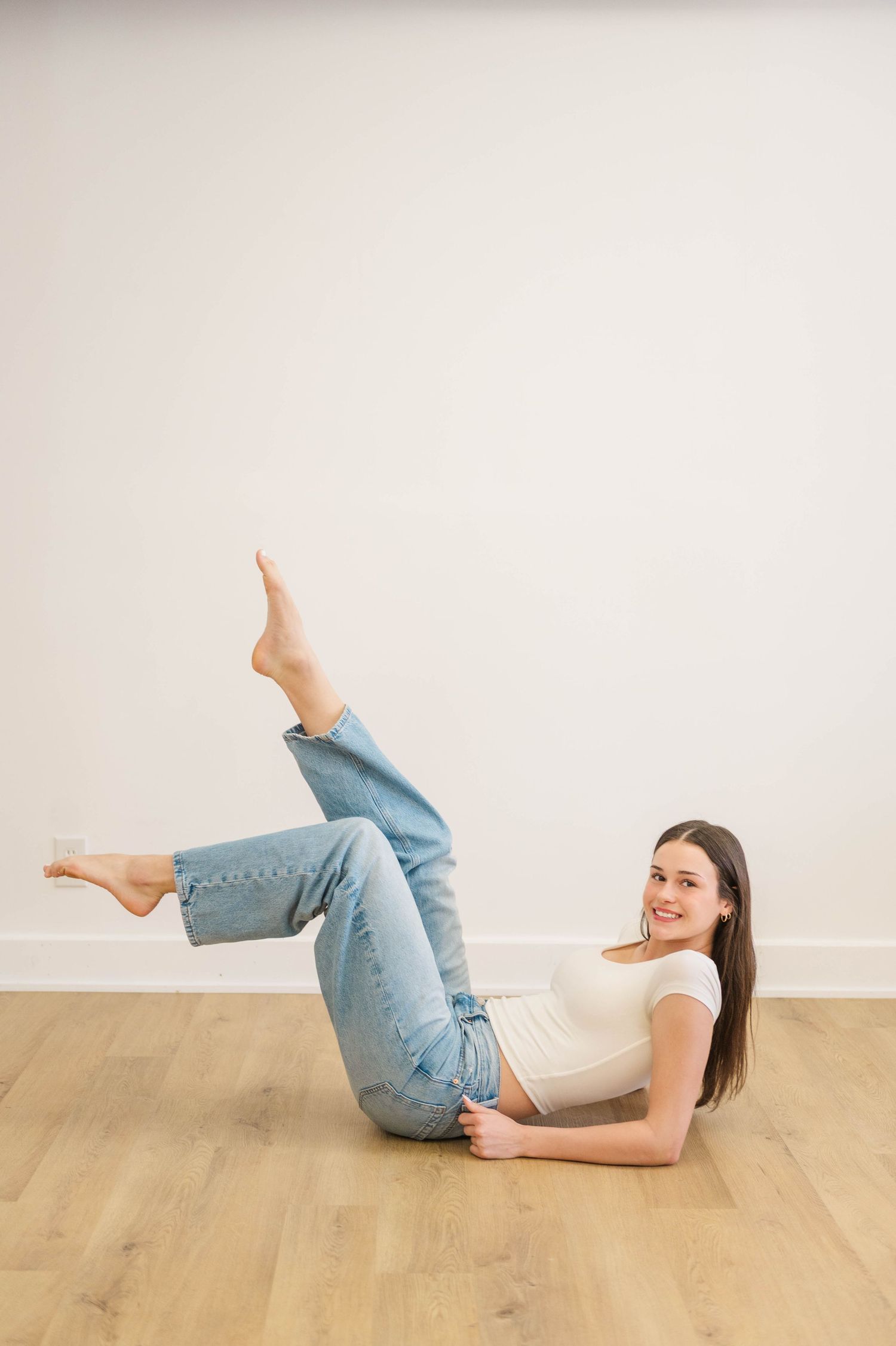 Brunette woman in white tee and light jeans lying on floor with legs raised up.