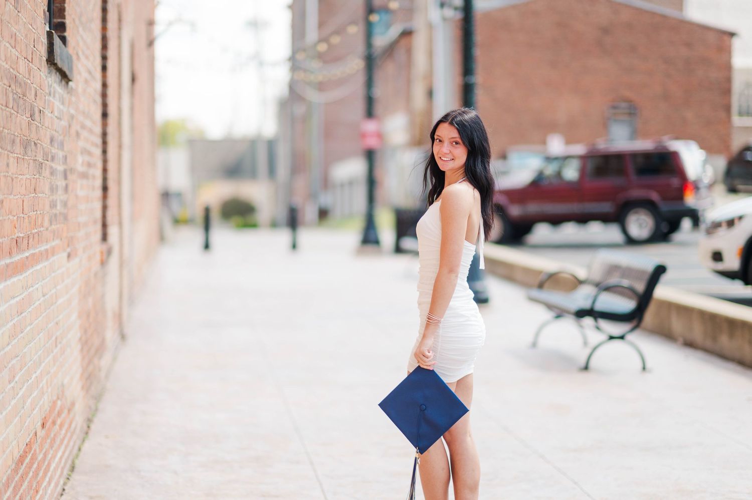Young woman in white dress carries navy graduation cap on sunny urban sidewalk.