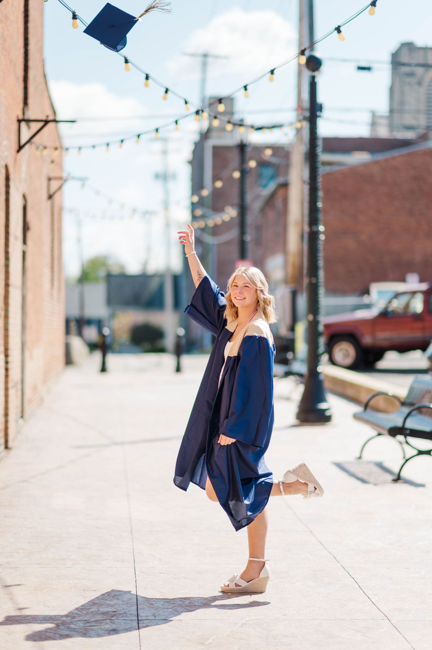 Smiling graduate in navy cap and gown tossing mortarboard in the air on a sunny urban sidewalk.