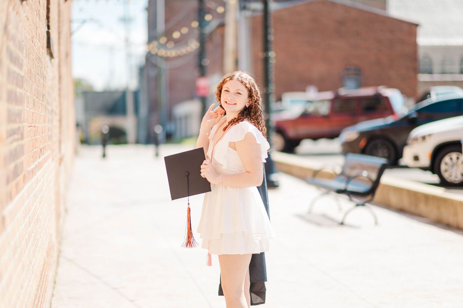 Happy graduate in white dress holding black cap on sunny city sidewalk.