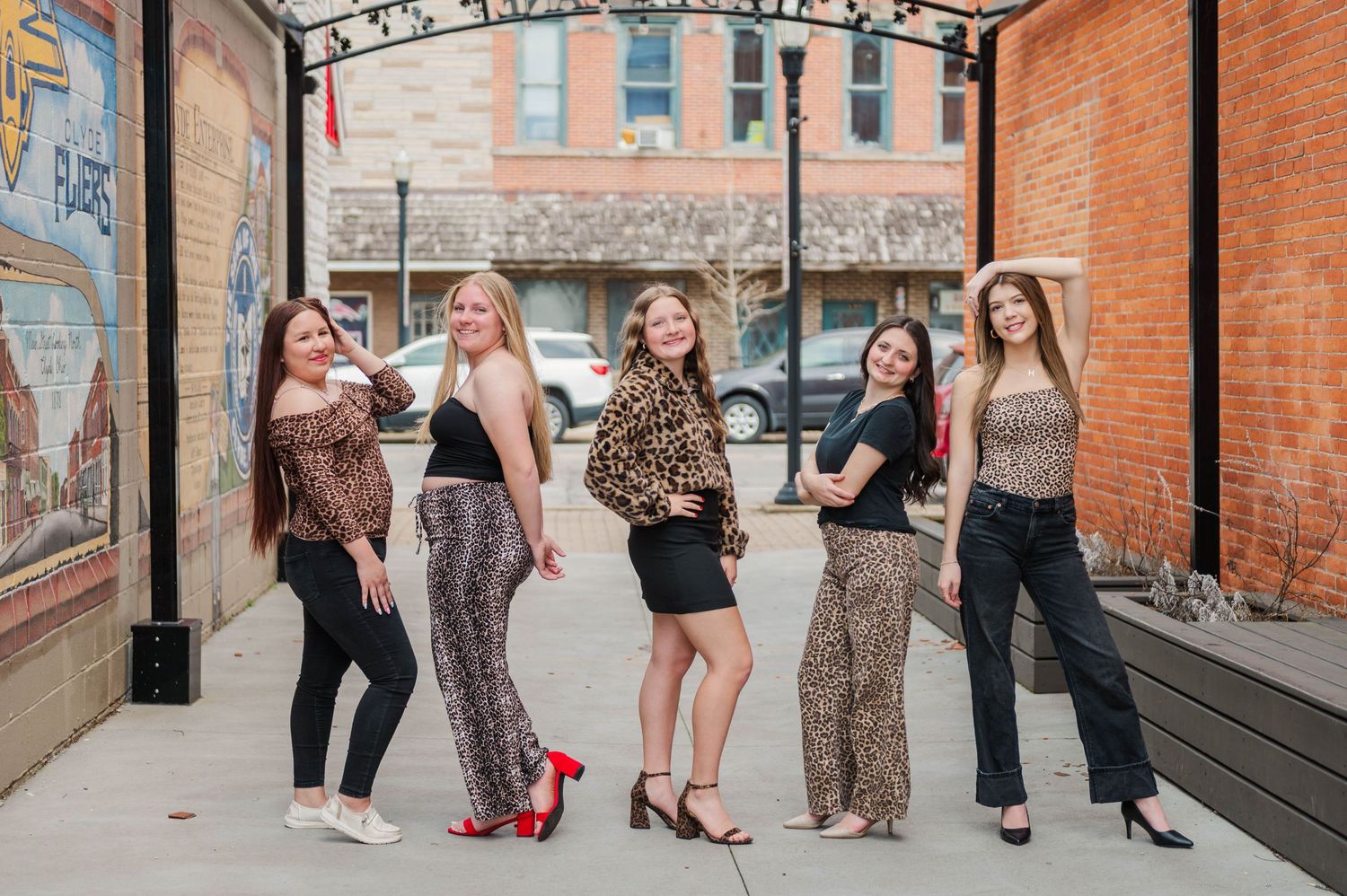 Five stylish women posing together on a sidewalk in front of a brick building with a mural.