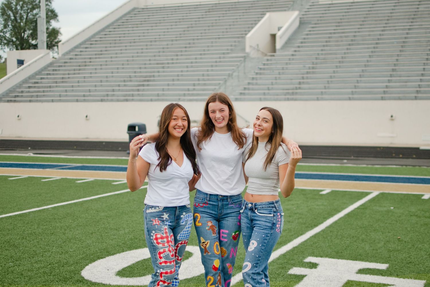 Three girls in white tops and colorful painted senior jeans take a selfie on a football field.