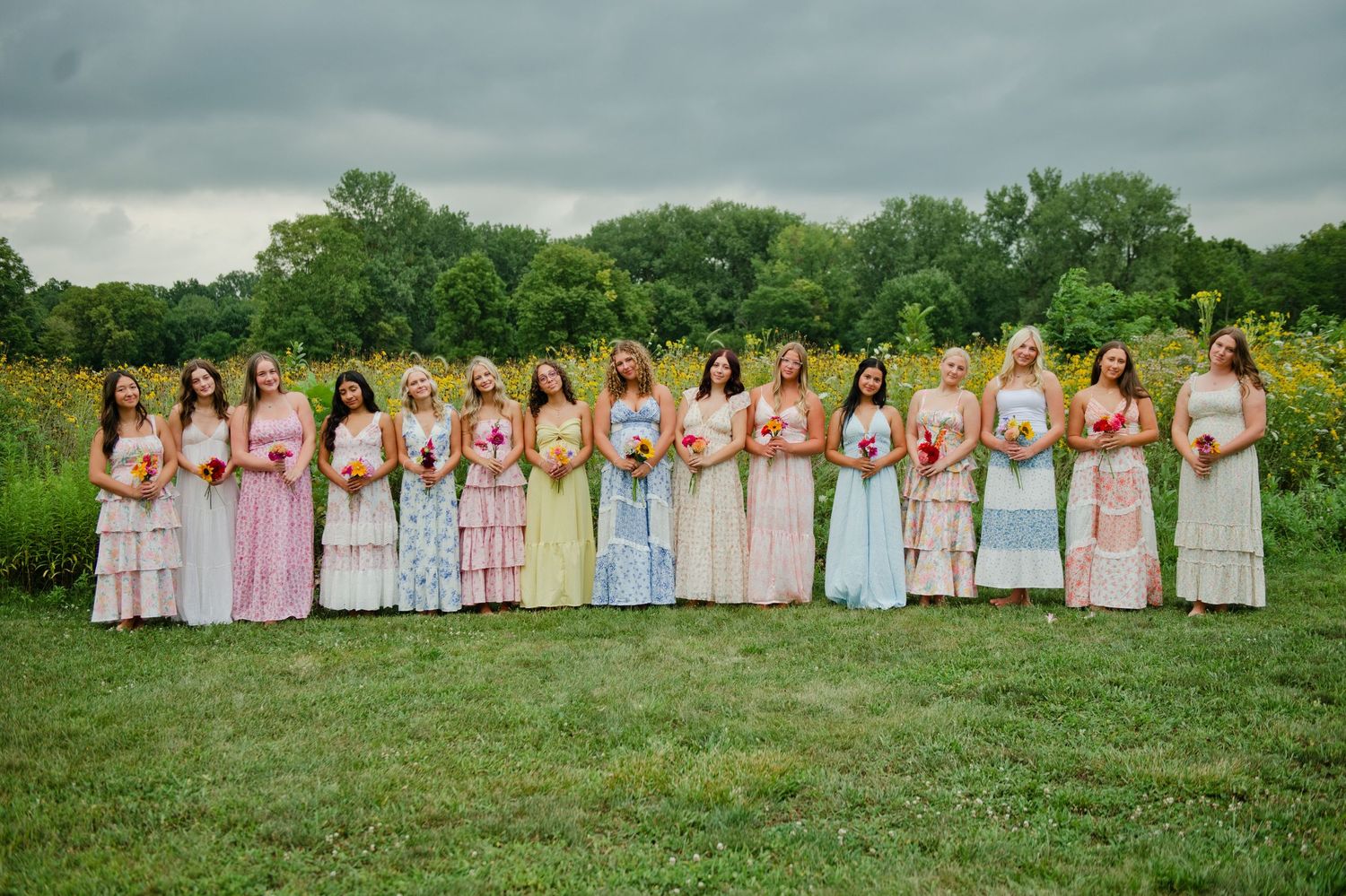 Large bridal party in mismatched pastel floral dresses holding bouquets in a sunflower field.