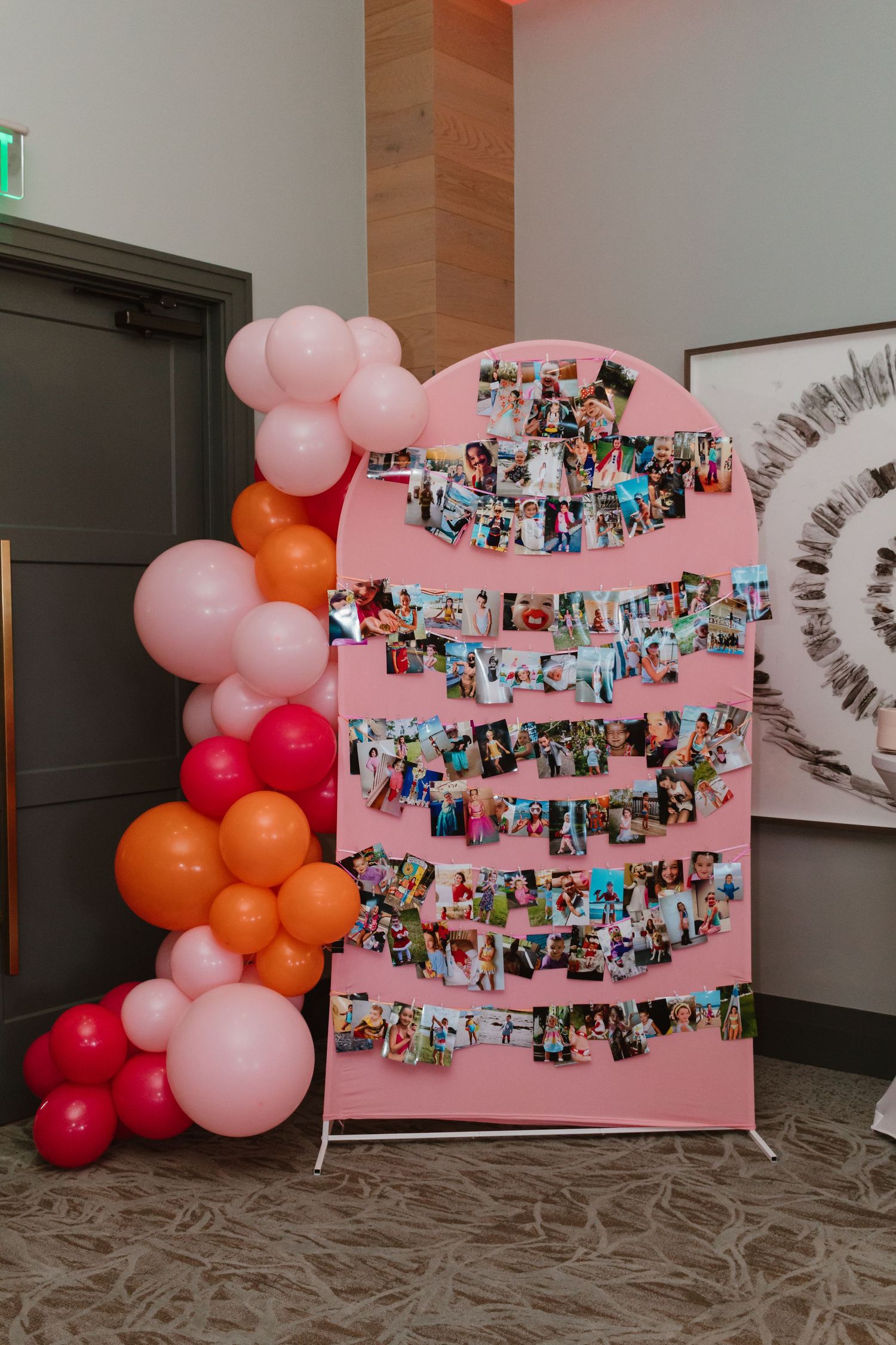 Pink photo display board covered in colorful printed photos surrounded by a pink, orange, and red balloon garland.