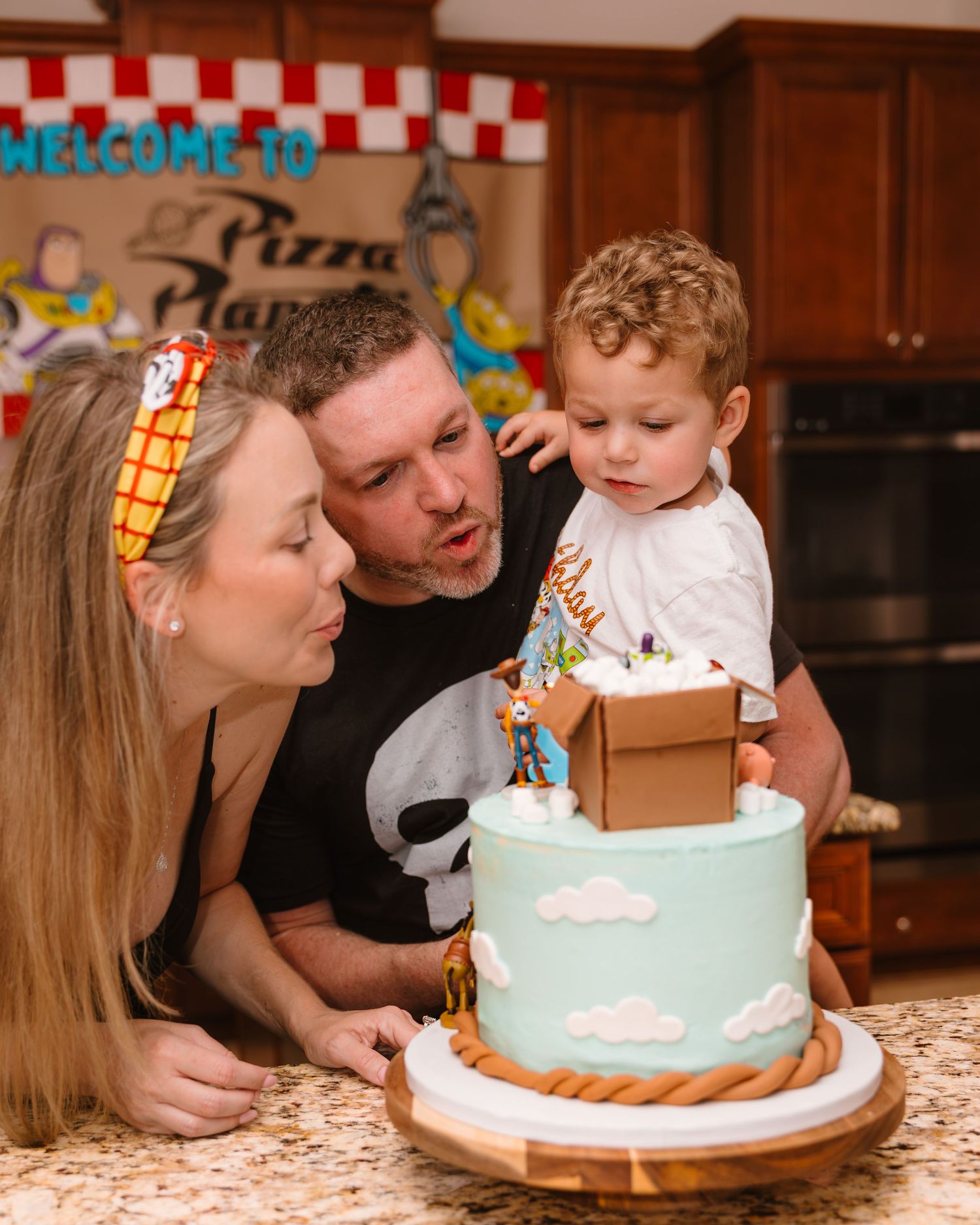 Parents help toddler blow out candles on Toy Story themed birthday cake at Pizza Planet party.
