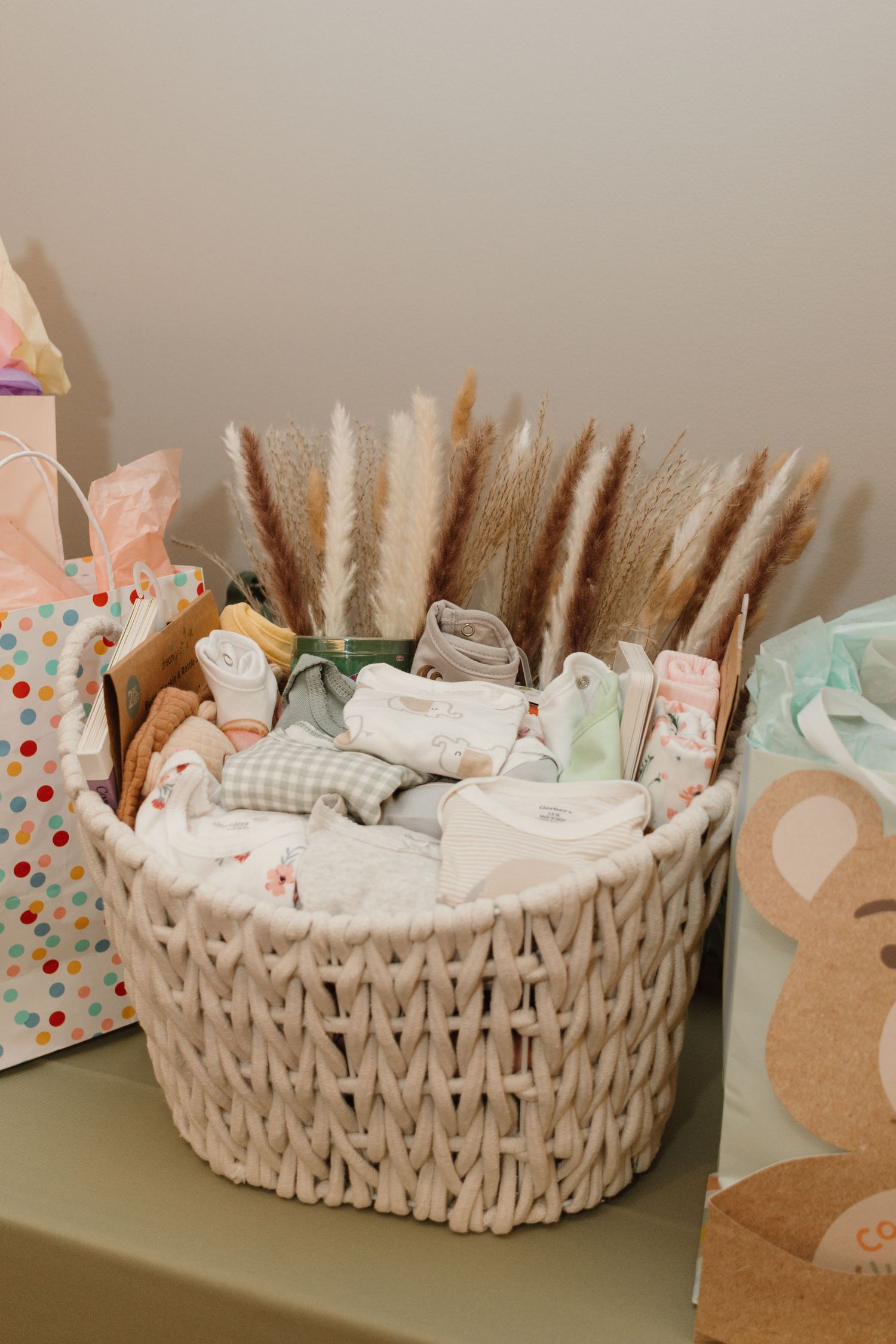 Woven white basket filled with folded baby clothes and dried pampas grass, surrounded by colorful gift bags.