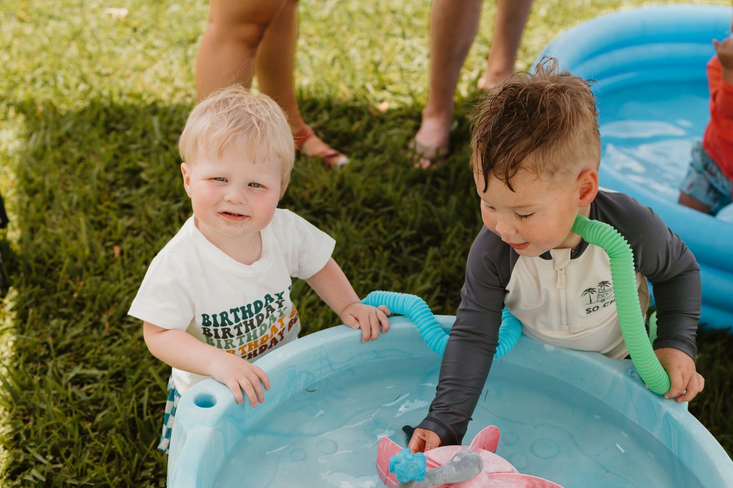 Two wet toddler boys playing together in blue inflatable pool at outdoor birthday party.