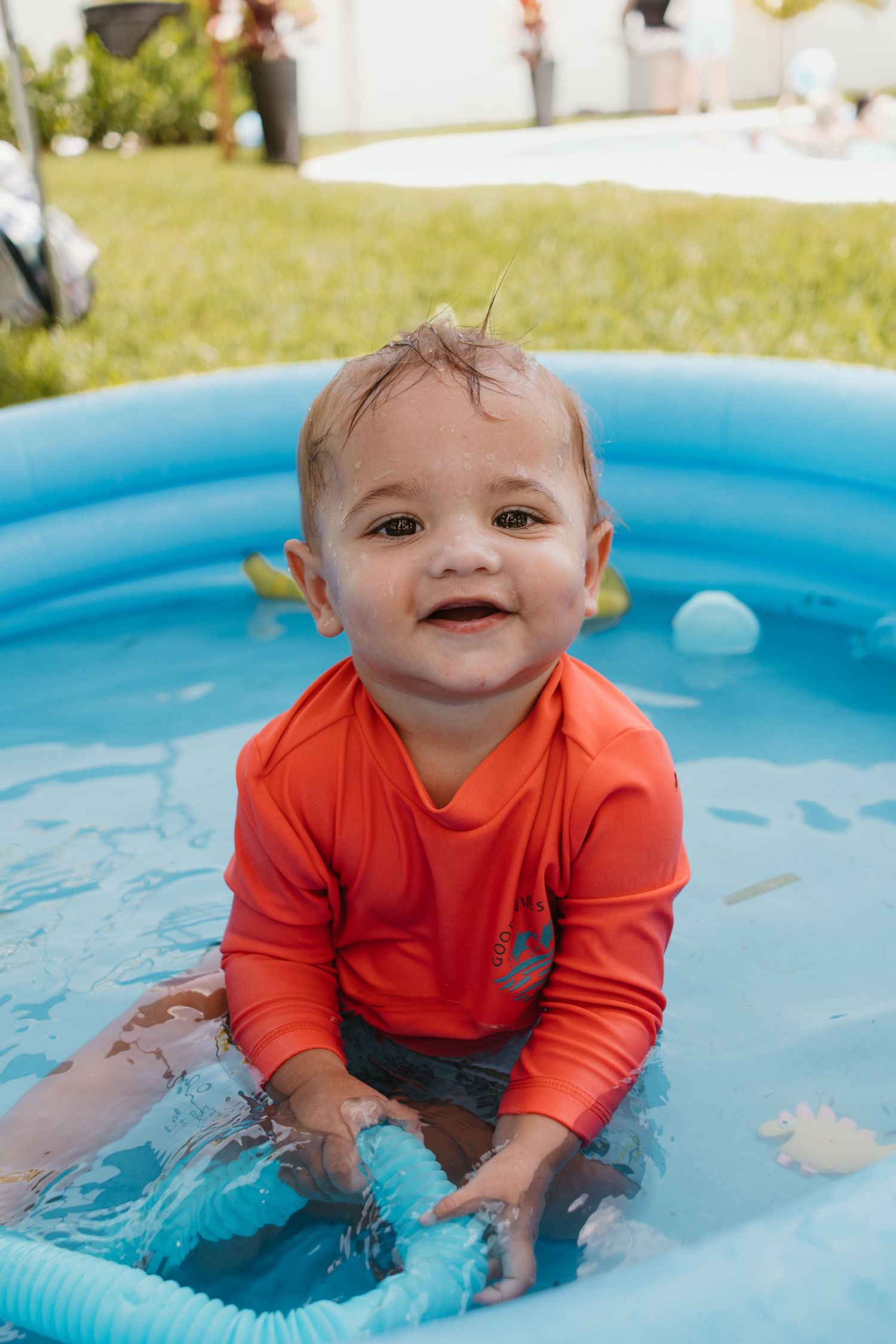 Happy baby in red rashguard splashing in blue inflatable pool at outdoor birthday party.