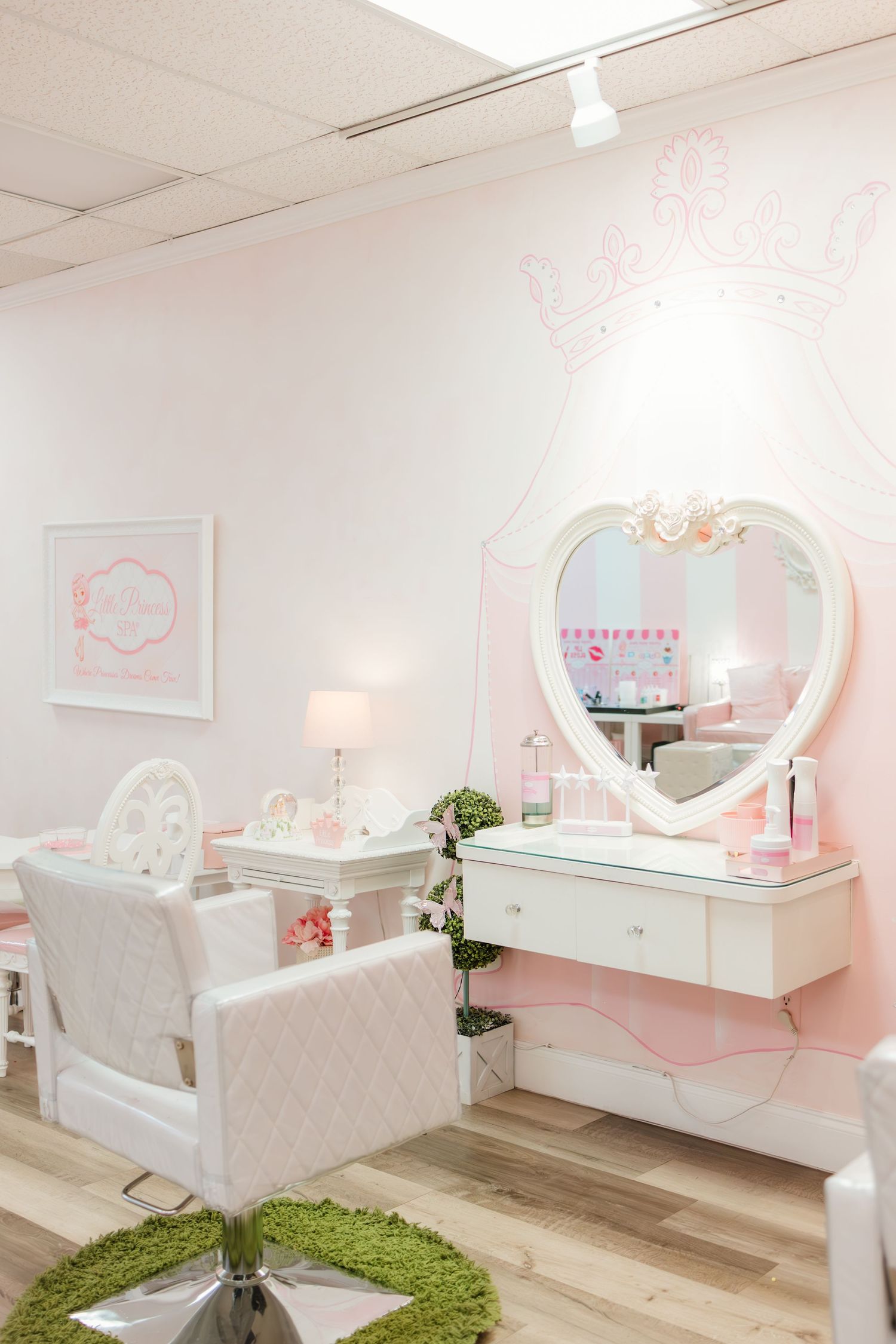 Pink princess salon with heart mirror, white vanity, tufted chair, and green rug on light wood floors.