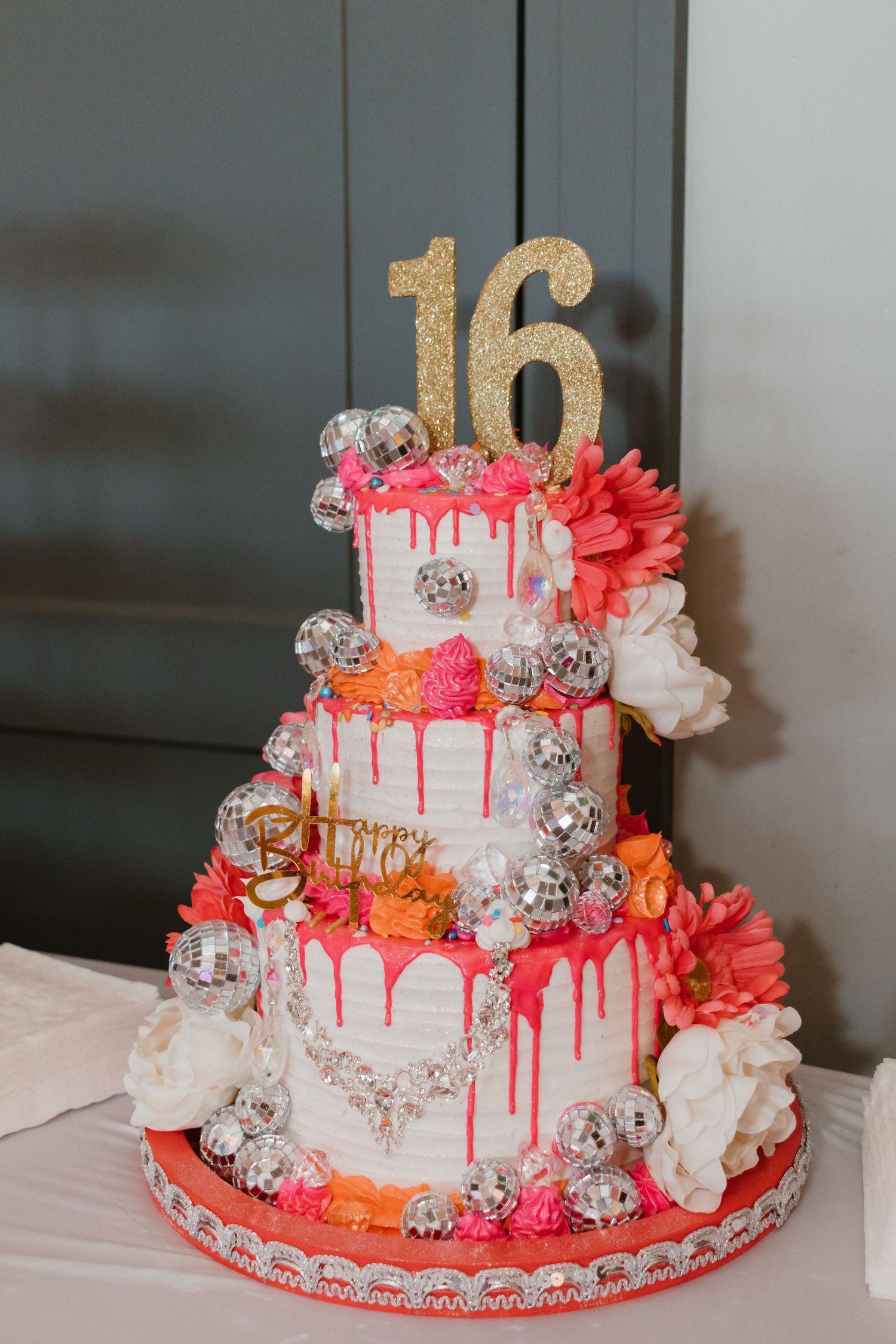 Three-tier white Sweet 16 birthday cake with pink drip frosting, disco balls, coral flowers, and gold number topper.