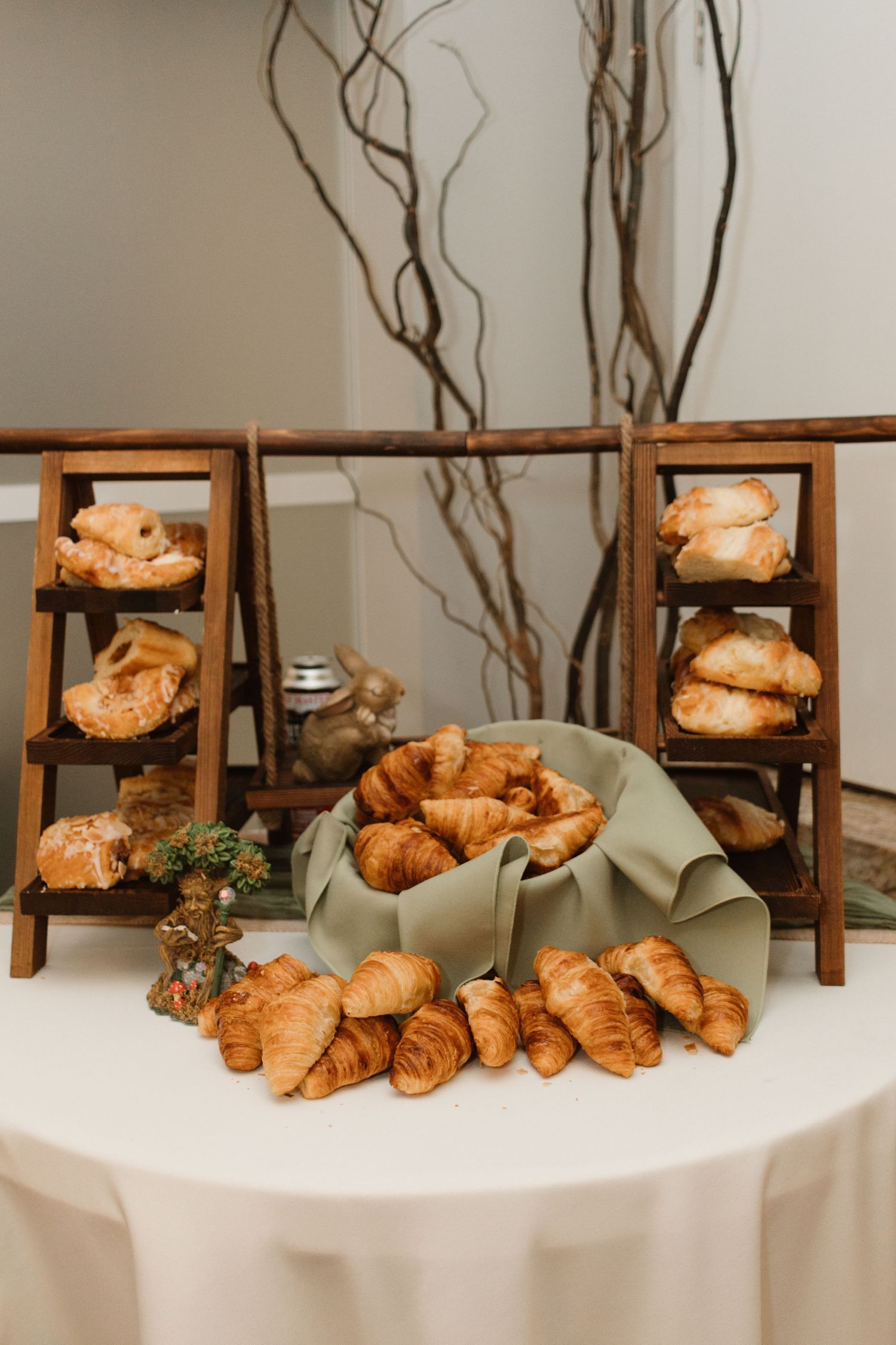 Elegant croissant display with wooden tiered stands and pastries arranged on a white tablecloth with rustic branch decor.